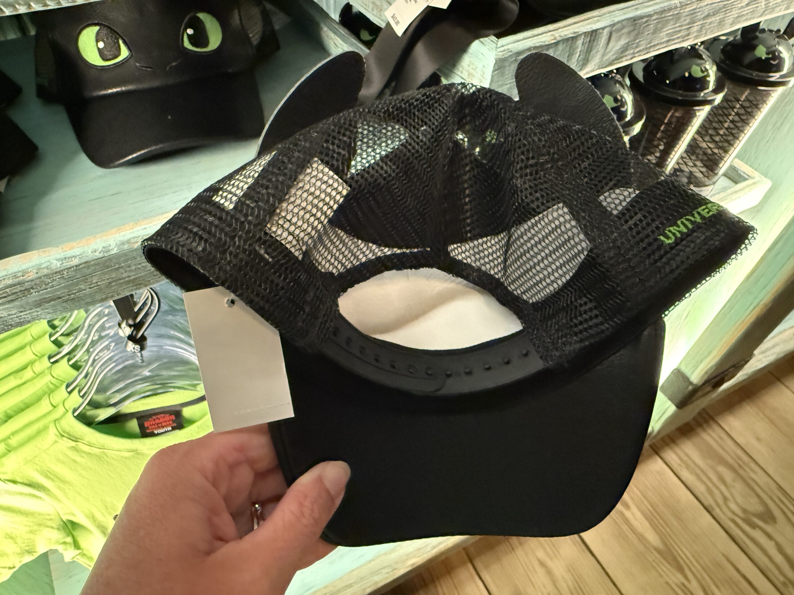A hand displays a black mesh cat-ear cap at the Spit Fyre Merch Kiosk in a Disney park, surrounded by themed souvenirs.