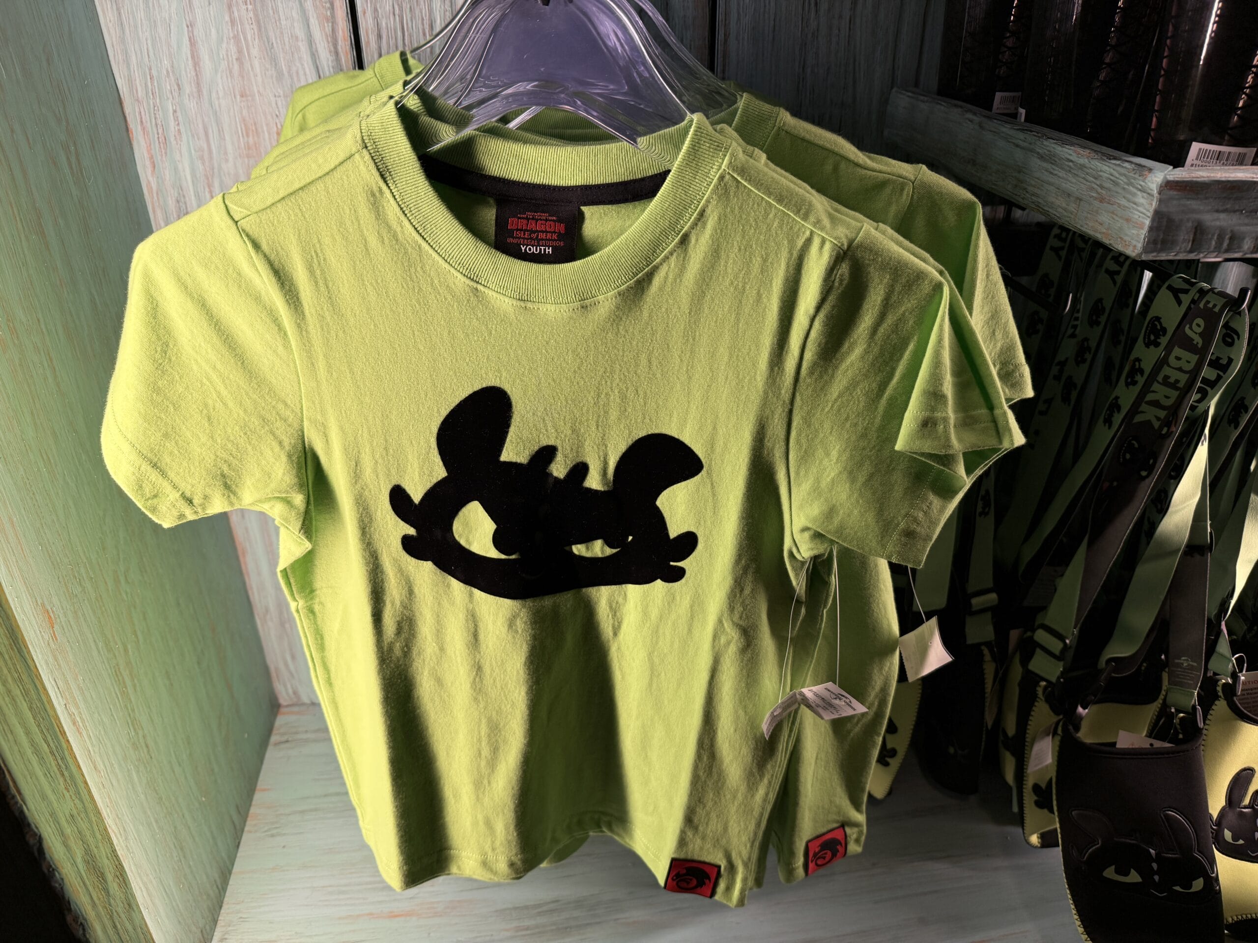 Bright green Spit Fyre kids' T-shirt with a bold cartoon face, seen at a Disney Park merch stand near iconic castle-themed displays.