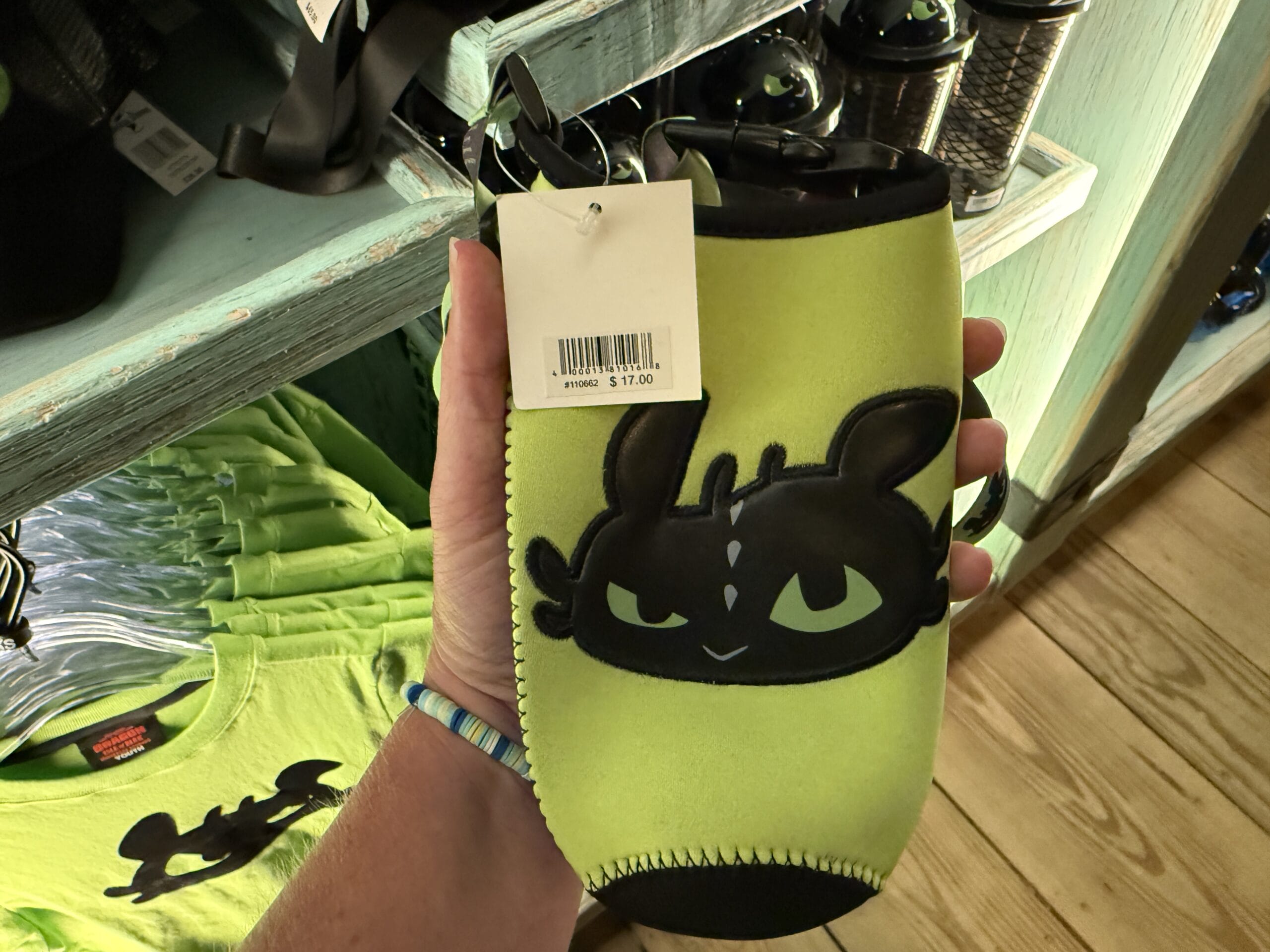 A hand displays a green can cooler with Toothless at the Spit Fyre kiosk for $17—no Disney park castle is visible here.