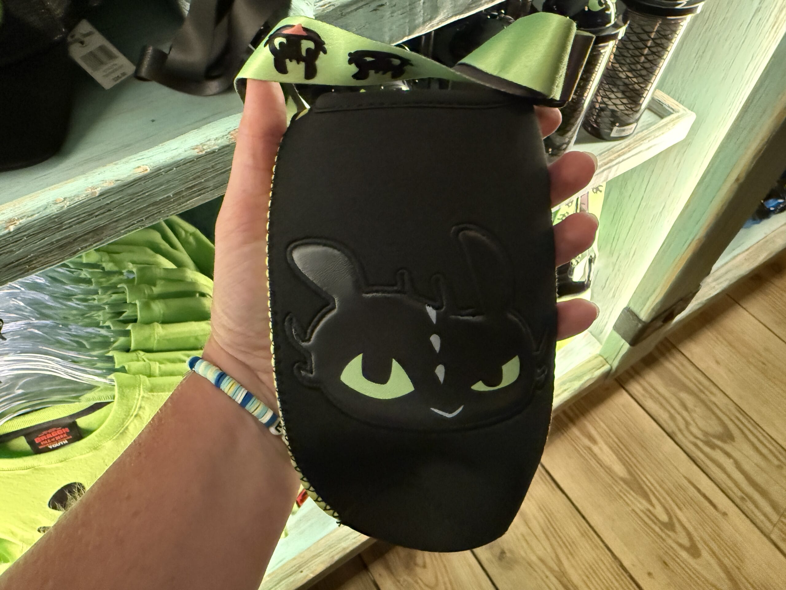 A hand displays Spit Fyre merch—a black Toothless pouch with green strap, reminiscent of merch sold in Disney park kiosks.
