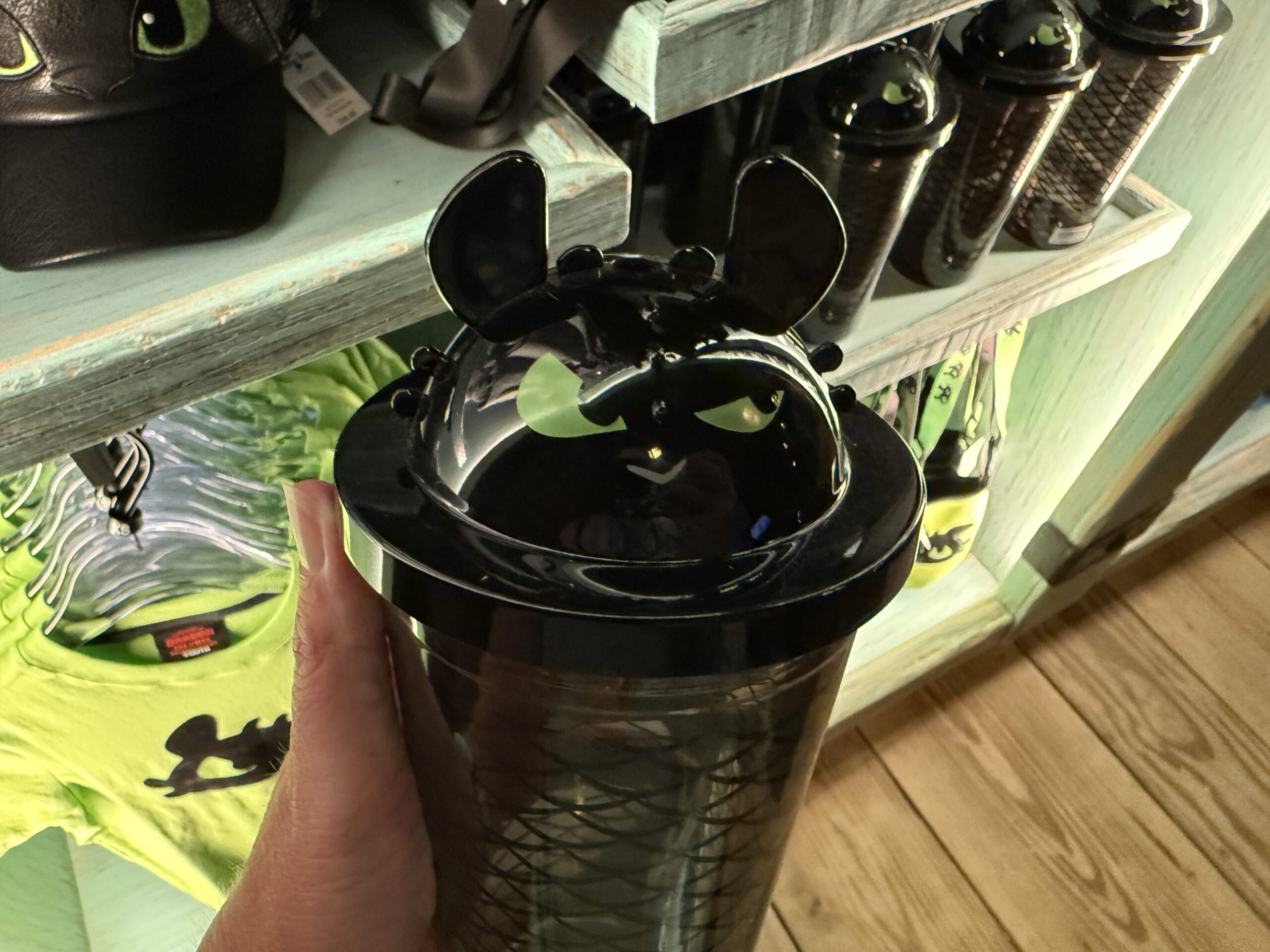 A hand displays Spit Fyre merch—a black tumbler with dragon face and ears—at a Disneyland park kiosk, no castle in view.