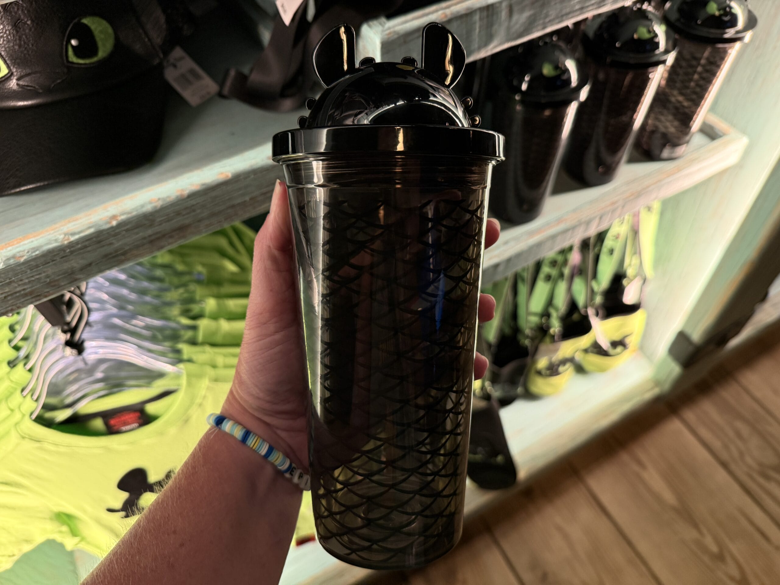 A hand holds a Spit Fyre black dragon-scale cup with green eyes, shown among themed merchandise at a Disney park kiosk shelf.