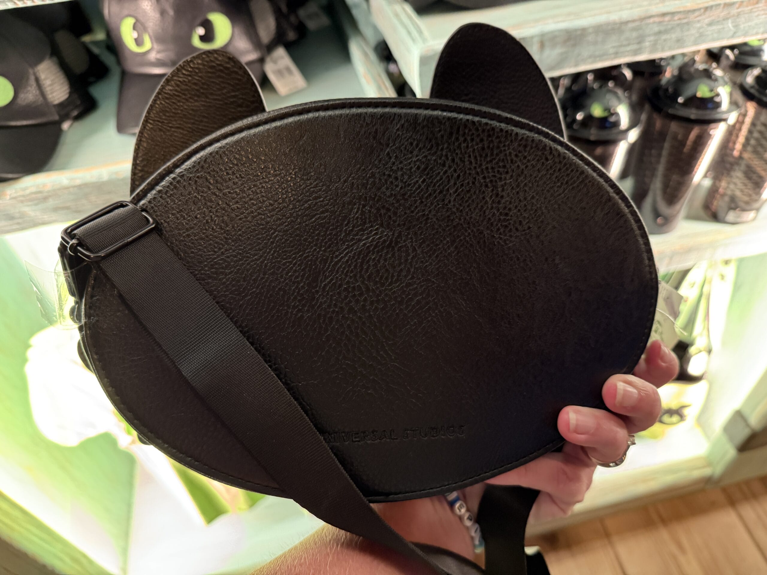 A hand displays a black cat-shaped bag with pointed ears at a Spit Fyre stand packed with feline Disney-themed merchandise.