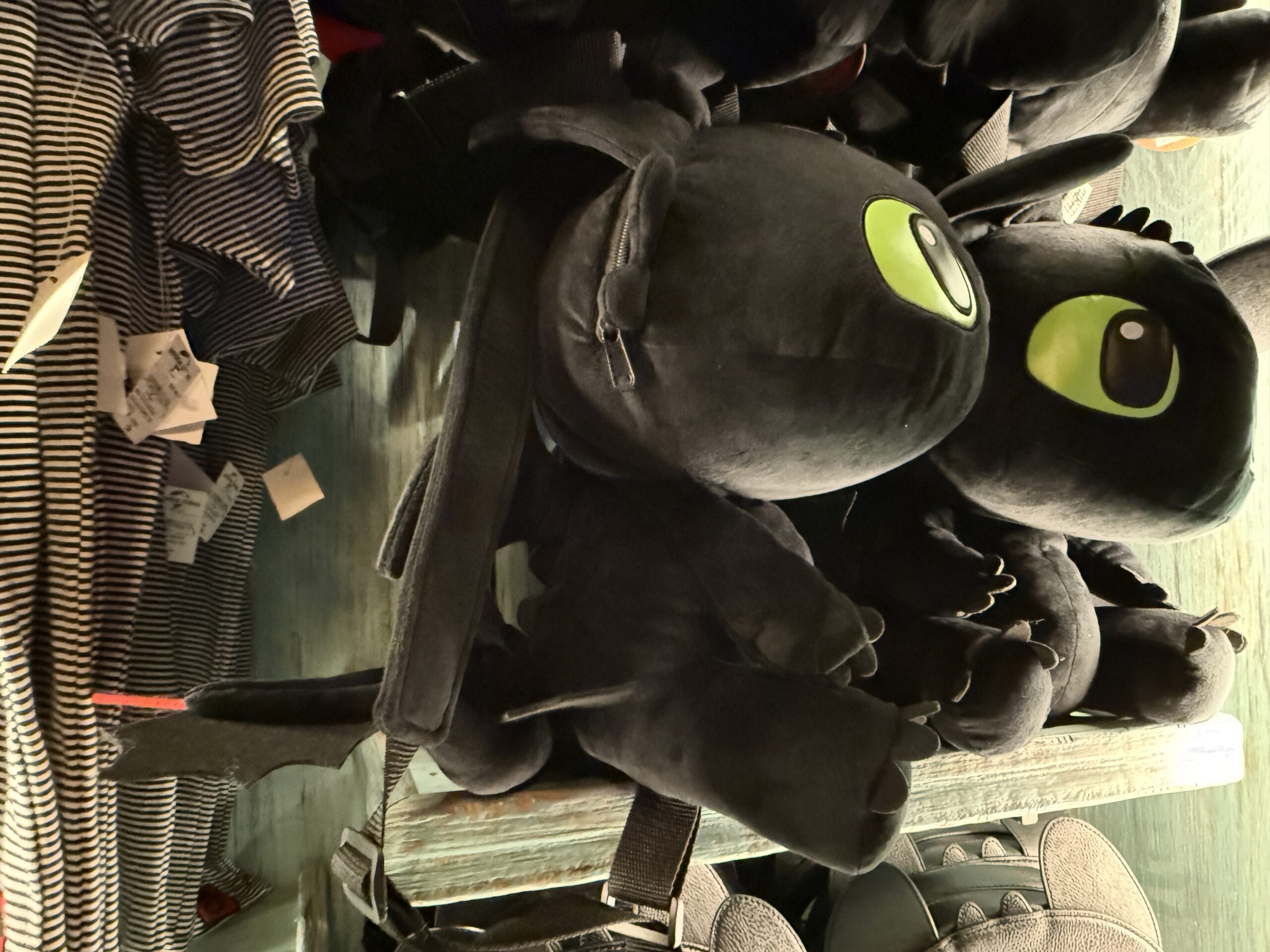 A plush Spit Fyre dragon toy with bright green eyes sits on a shelf beside striped merch, reminiscent of Disney park displays.