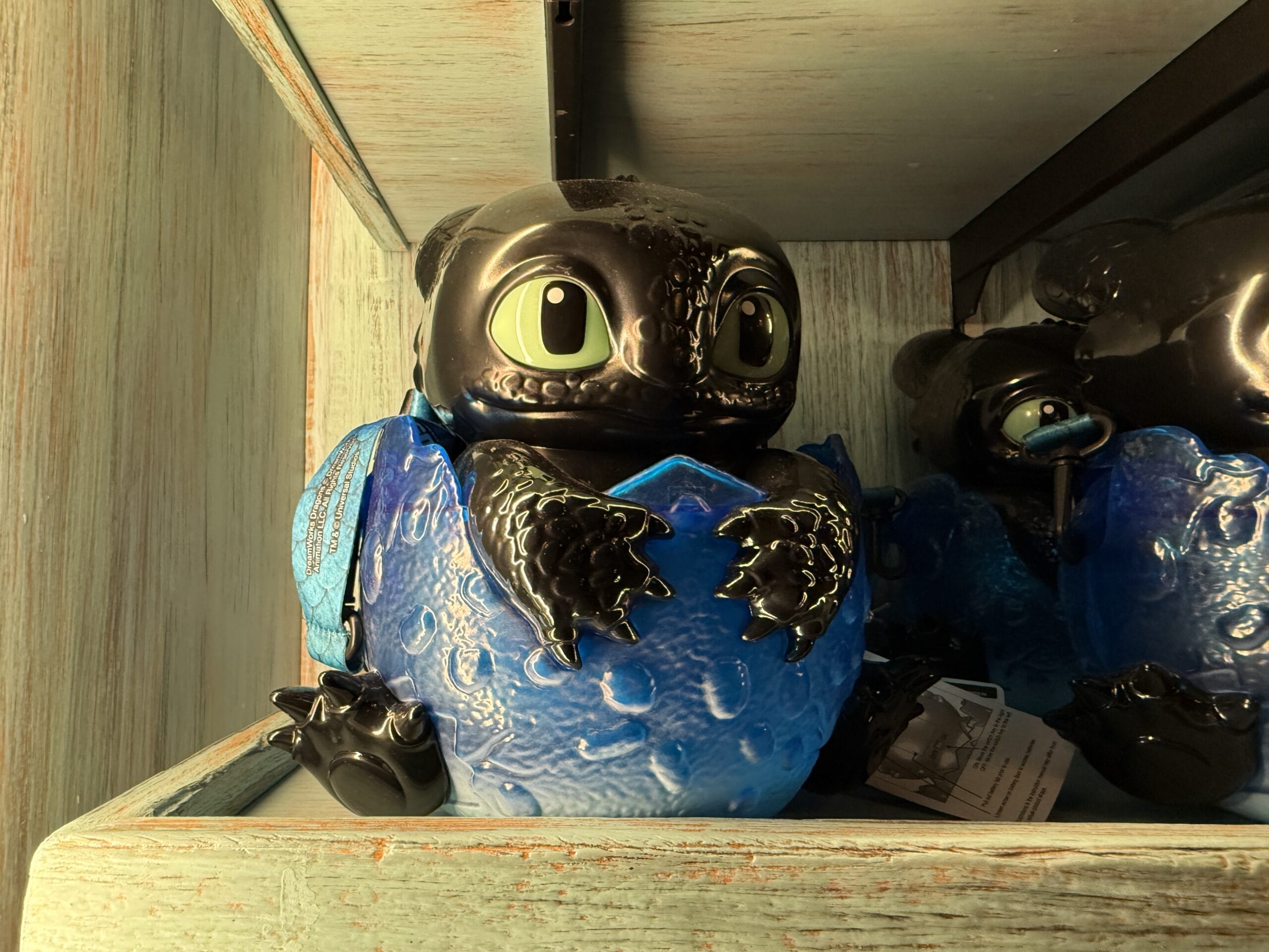 A Spit Fyre toy dragon with green eyes sits in a blue egg on a Disney park kiosk shelf with other merchandise nearby.