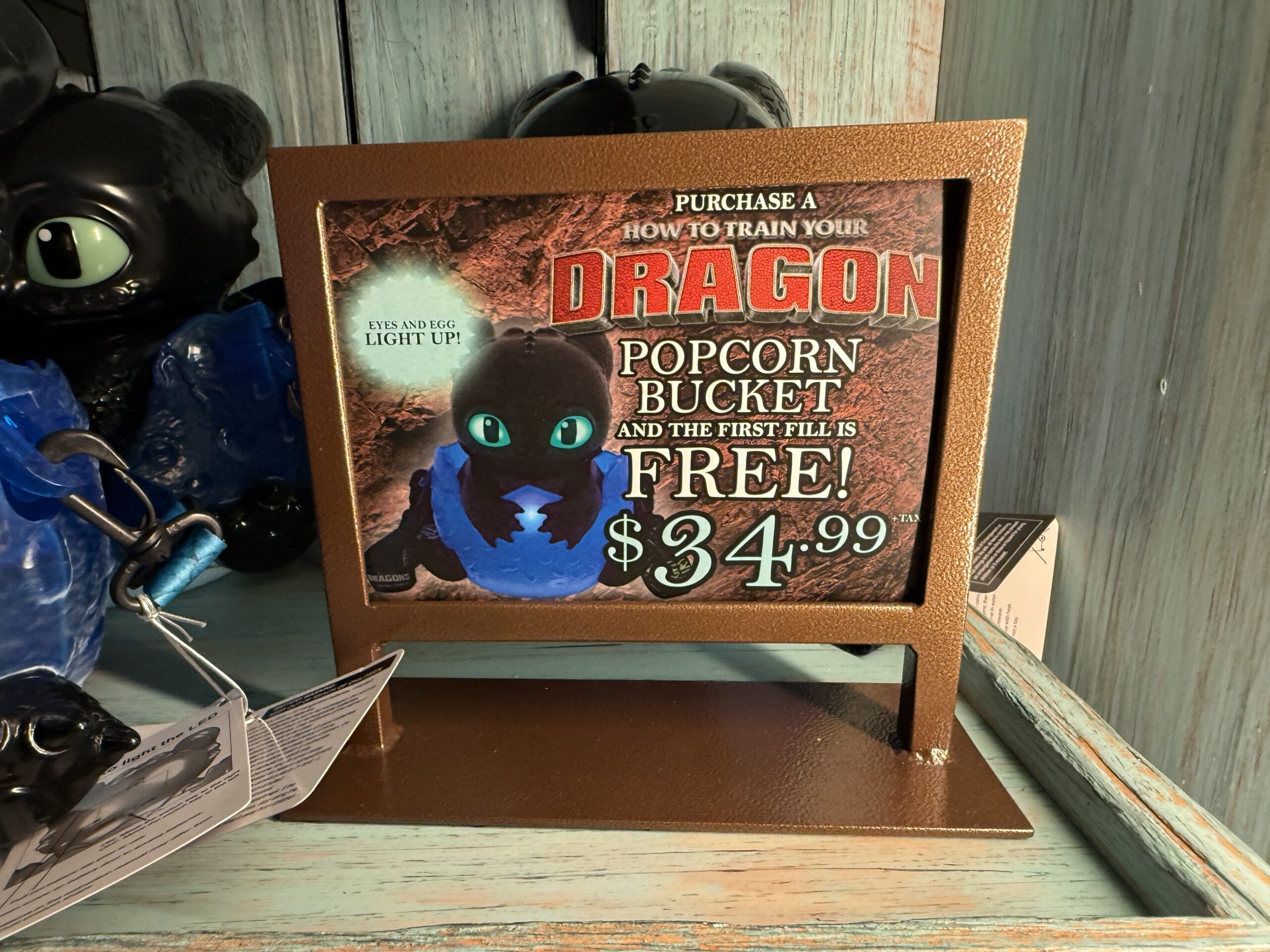 At a theme park kiosk, a Spit Fyre How to Train Your Dragon popcorn bucket is advertised for $34.99, first fill included.