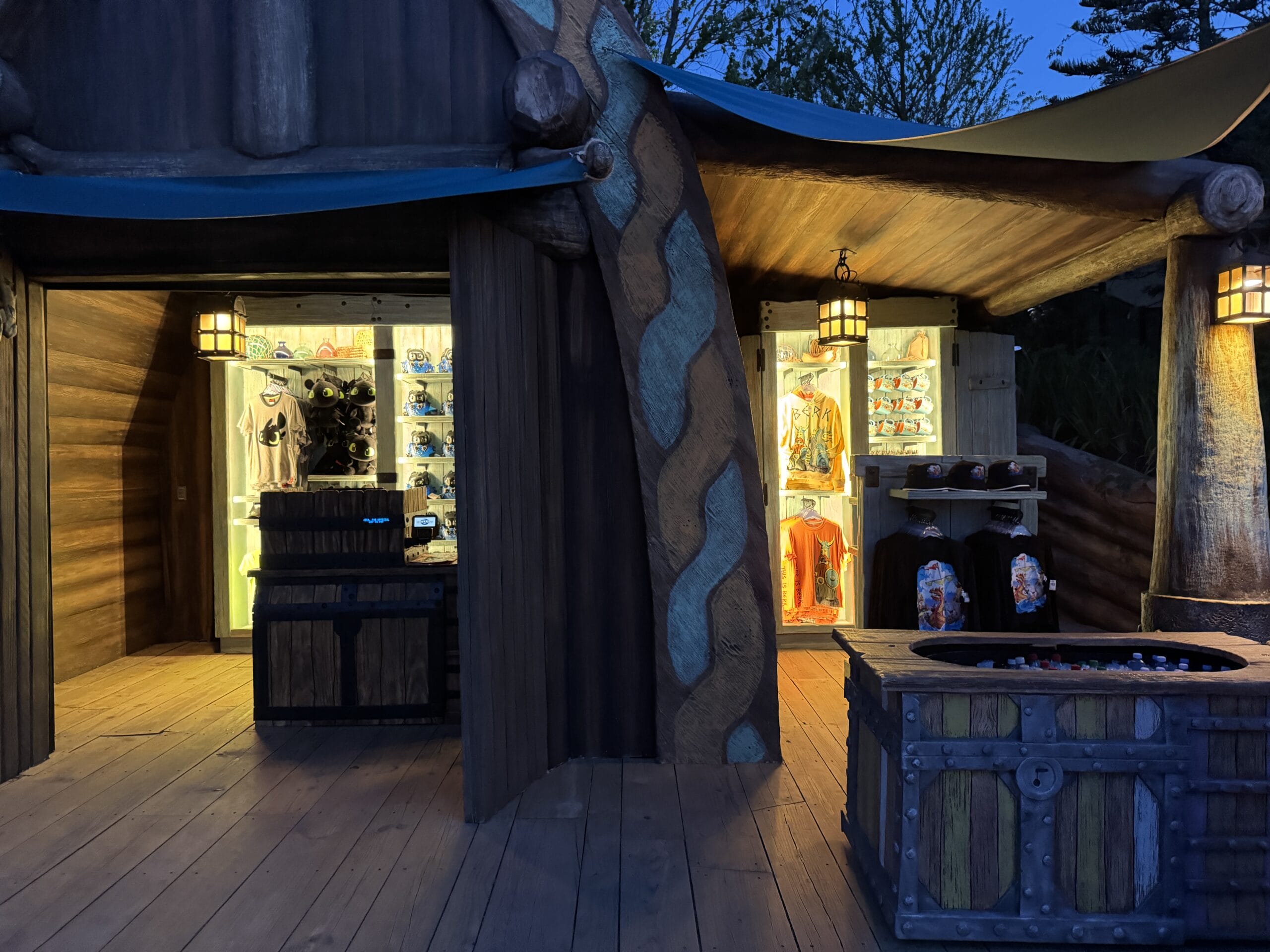 At dusk, a wooden Spit Fyre kiosk glows with warm lights, selling Disney park souvenirs, apparel, plush toys, and collectibles.