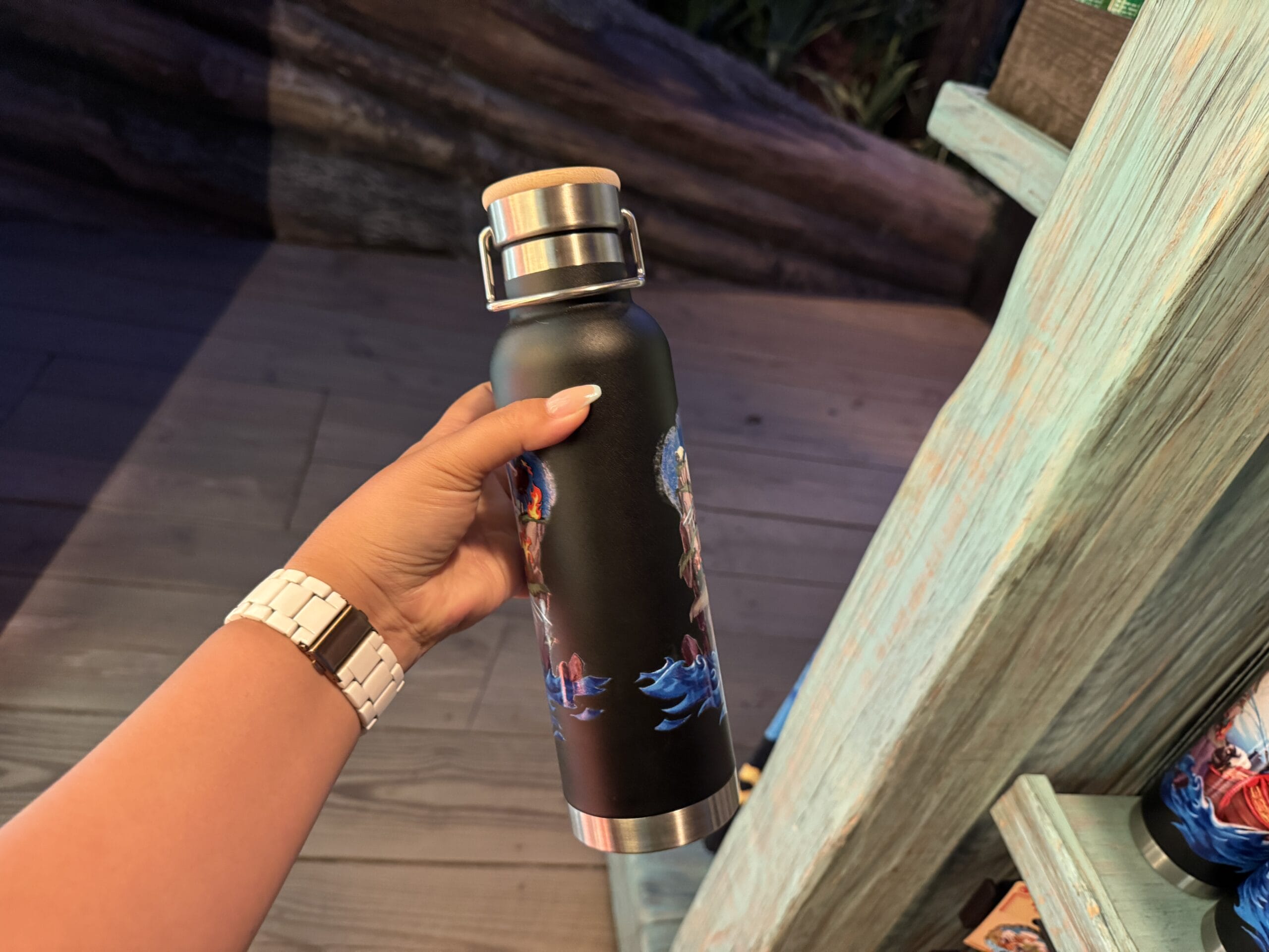 A hand holds a black stainless steel water bottle with colorful Disney-inspired designs near a light blue wooden shelf, reminiscent of park decor.