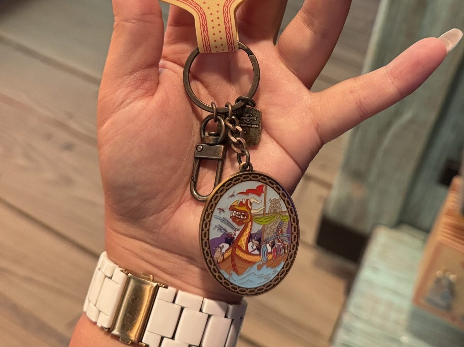 A hand holds a "Dragon: Isle of Berk" Spit Fyre keychain with a dragon design at a merch kiosk, not a Disney castle charm.