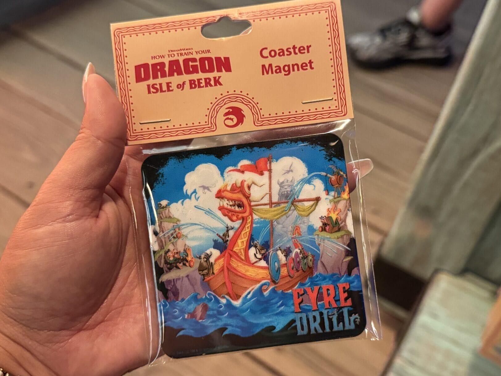 Hand holding a "Dragon Isle of Berk" coaster magnet featuring a red dragon, Viking ship, and "Fire Drill" text—no Disney castle.