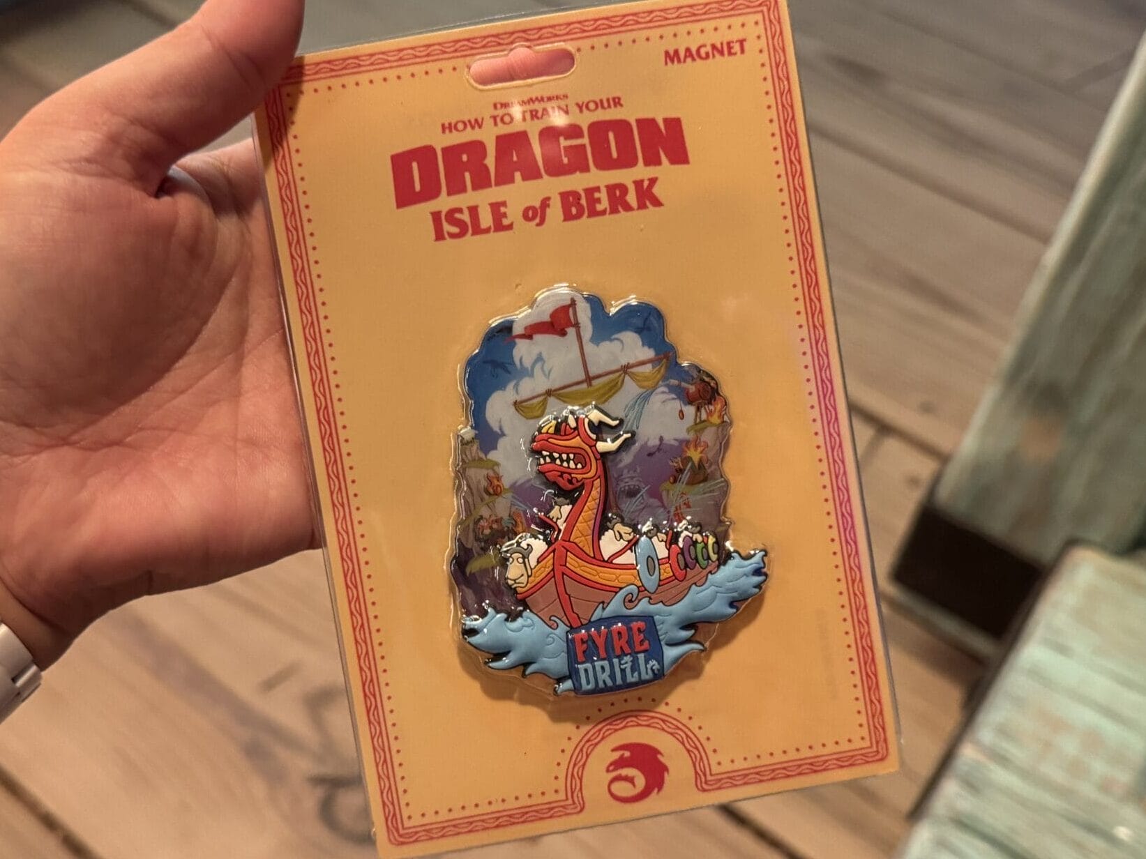 A hand holds a "How to Train Your Dragon: Isle of Berk" magnet with a dragon and "FYRE DRILL," not a Disney castle like Cinderella's.