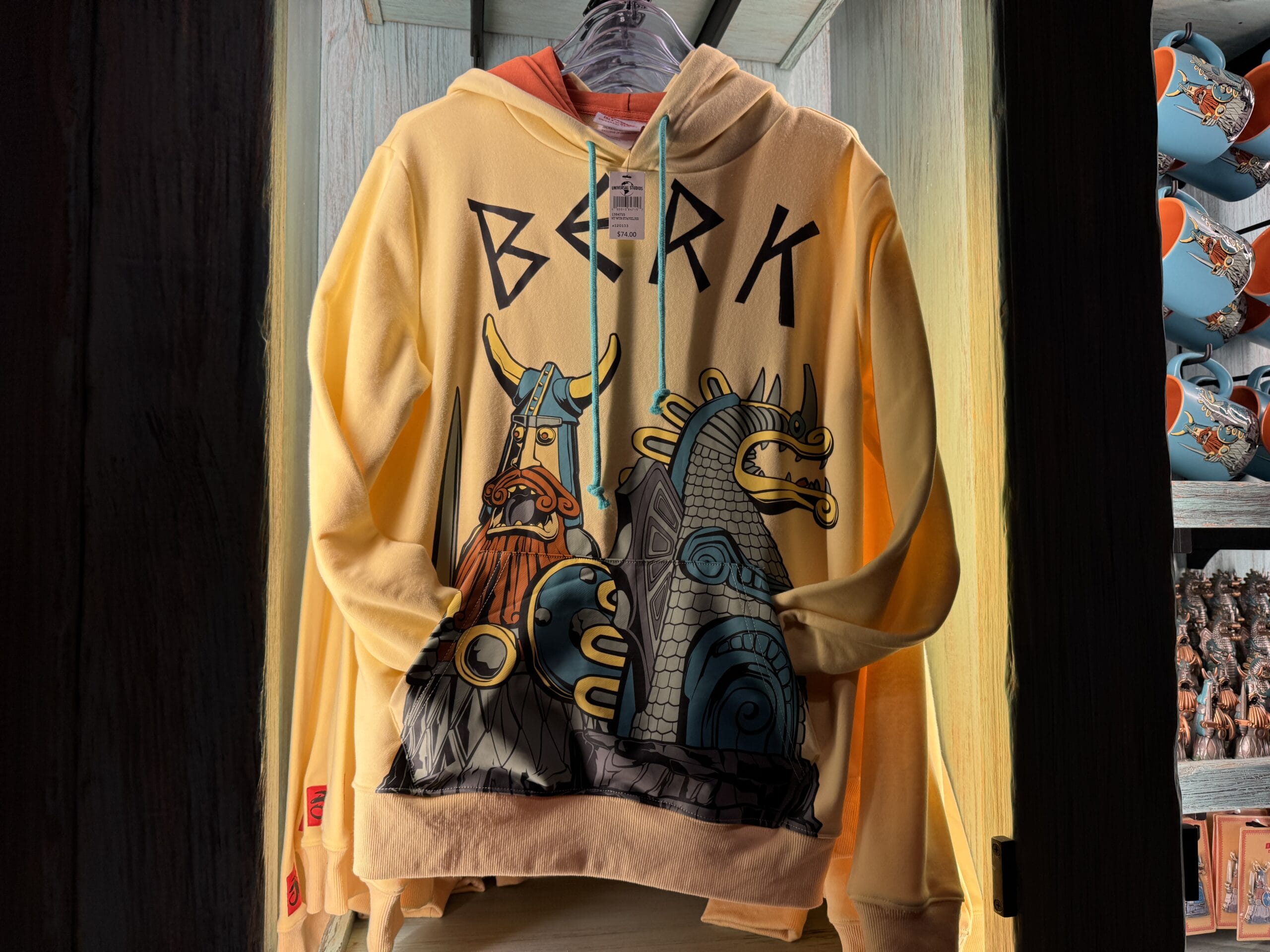 Yellow hoodie featuring Viking and dragon designs with "BERK" text, displayed at a theme park merch booth beside Spit Fyre mugs.