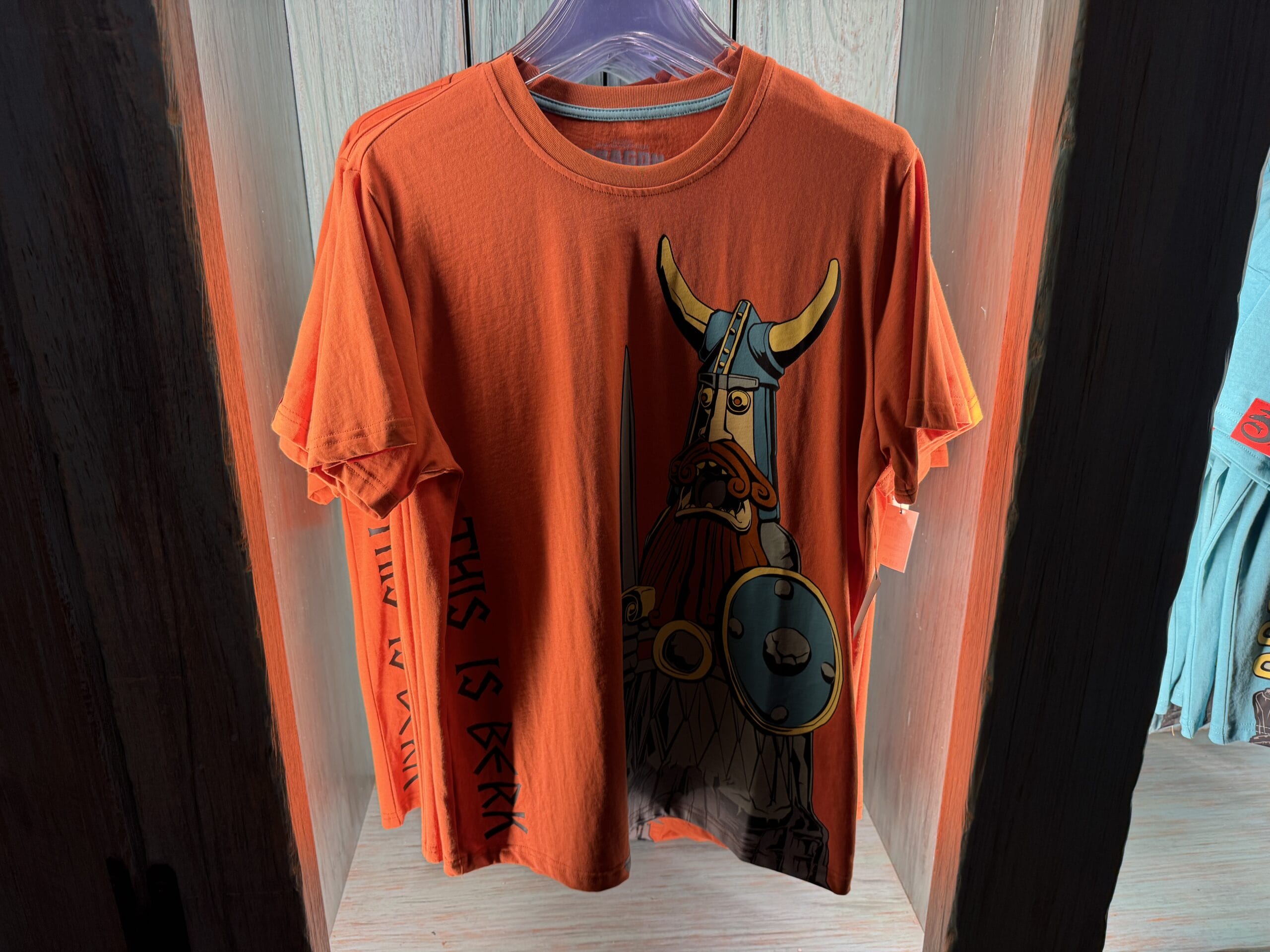 An orange T-shirt with a cartoon Viking warrior, sold at Spit Fyre kiosk, reads "THIS IS BERK"—not Disney Parks castle themed.