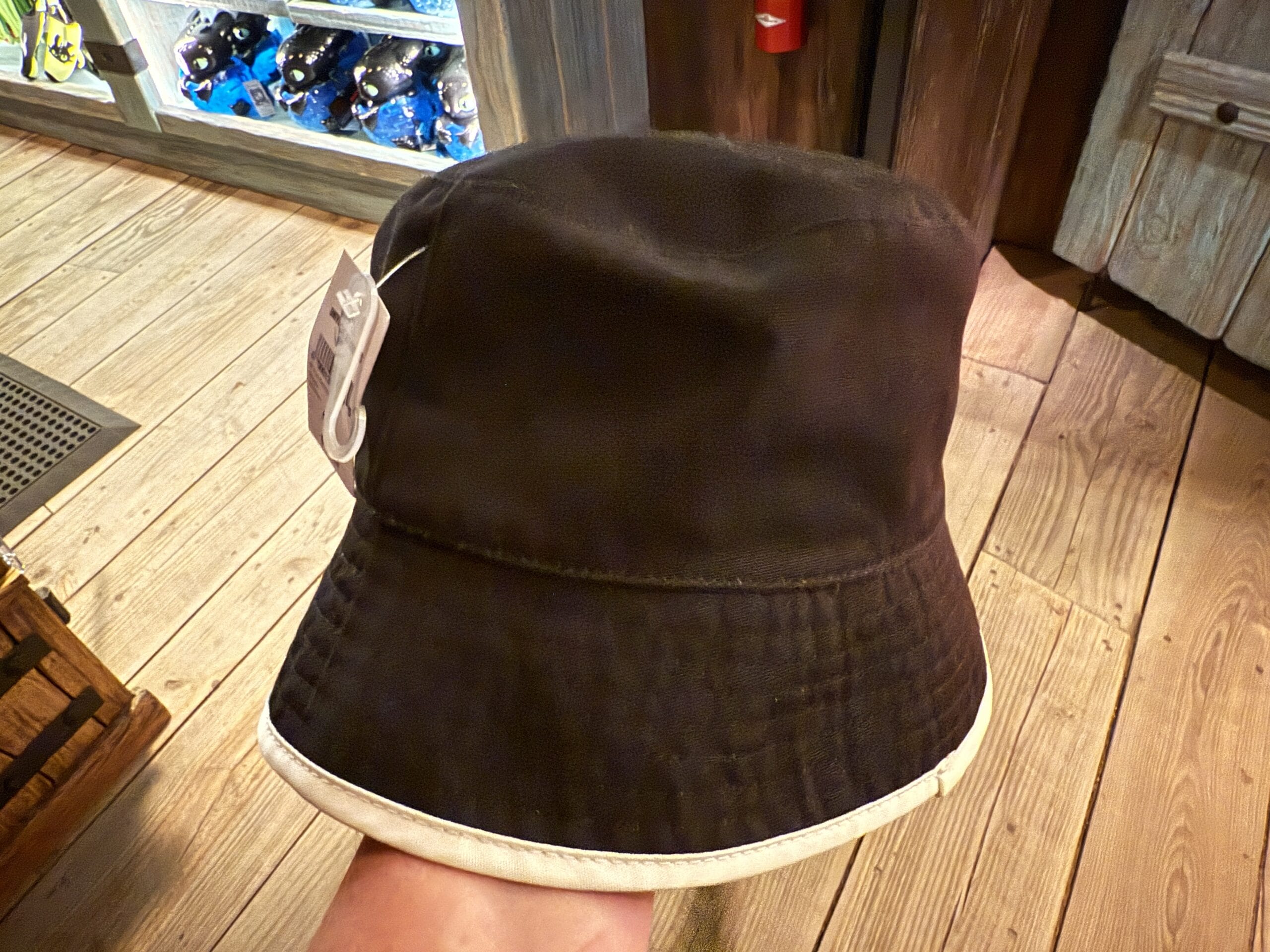 A black Spit Fyre bucket hat with a white rim is displayed in a kiosk—background resembles the shops along Main Street, U.S.A.