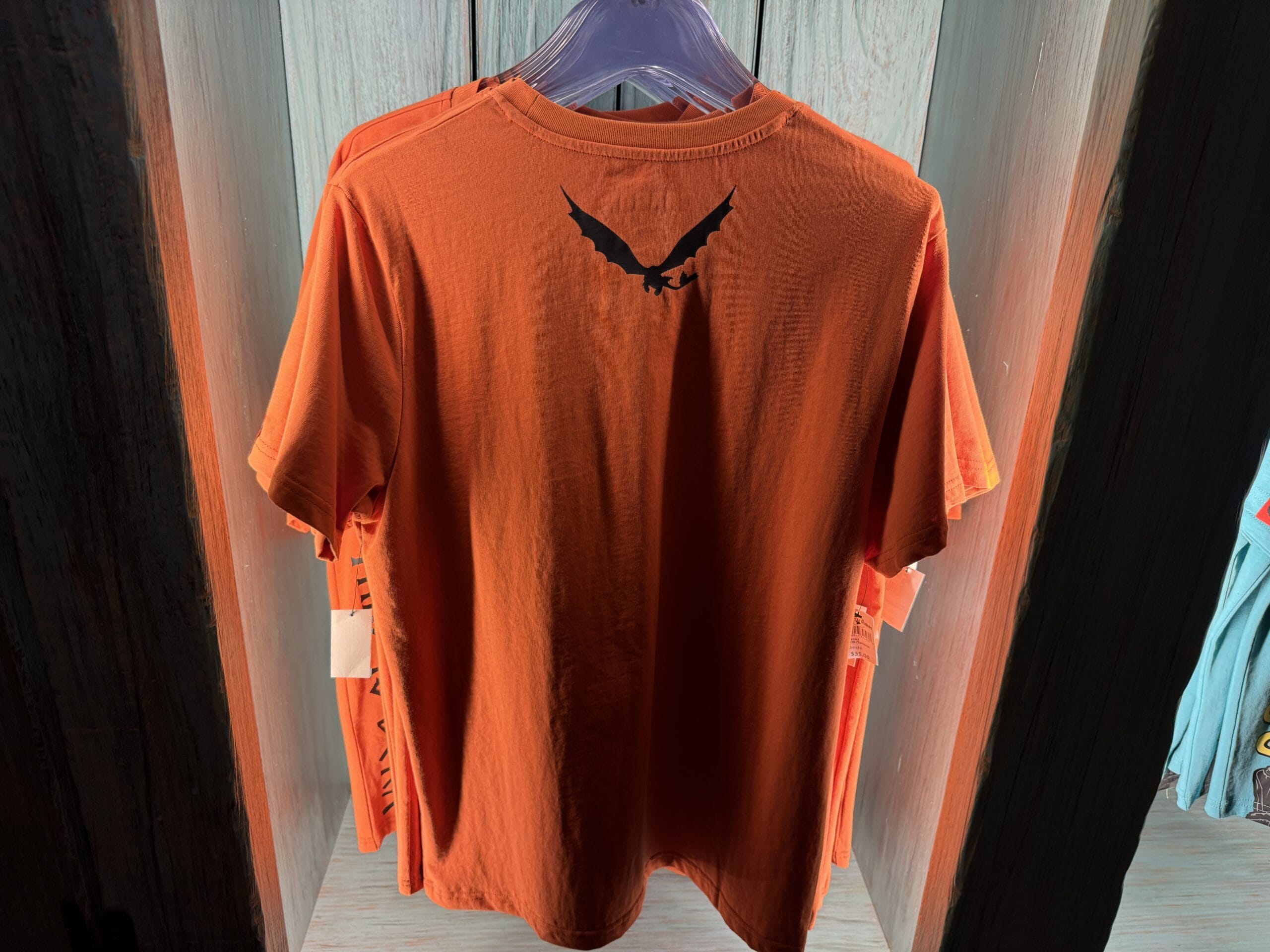 An orange Spit Fyre merchandise t-shirt hangs on a rack, featuring a black bird silhouette near the collar—a vibrant park souvenir.
