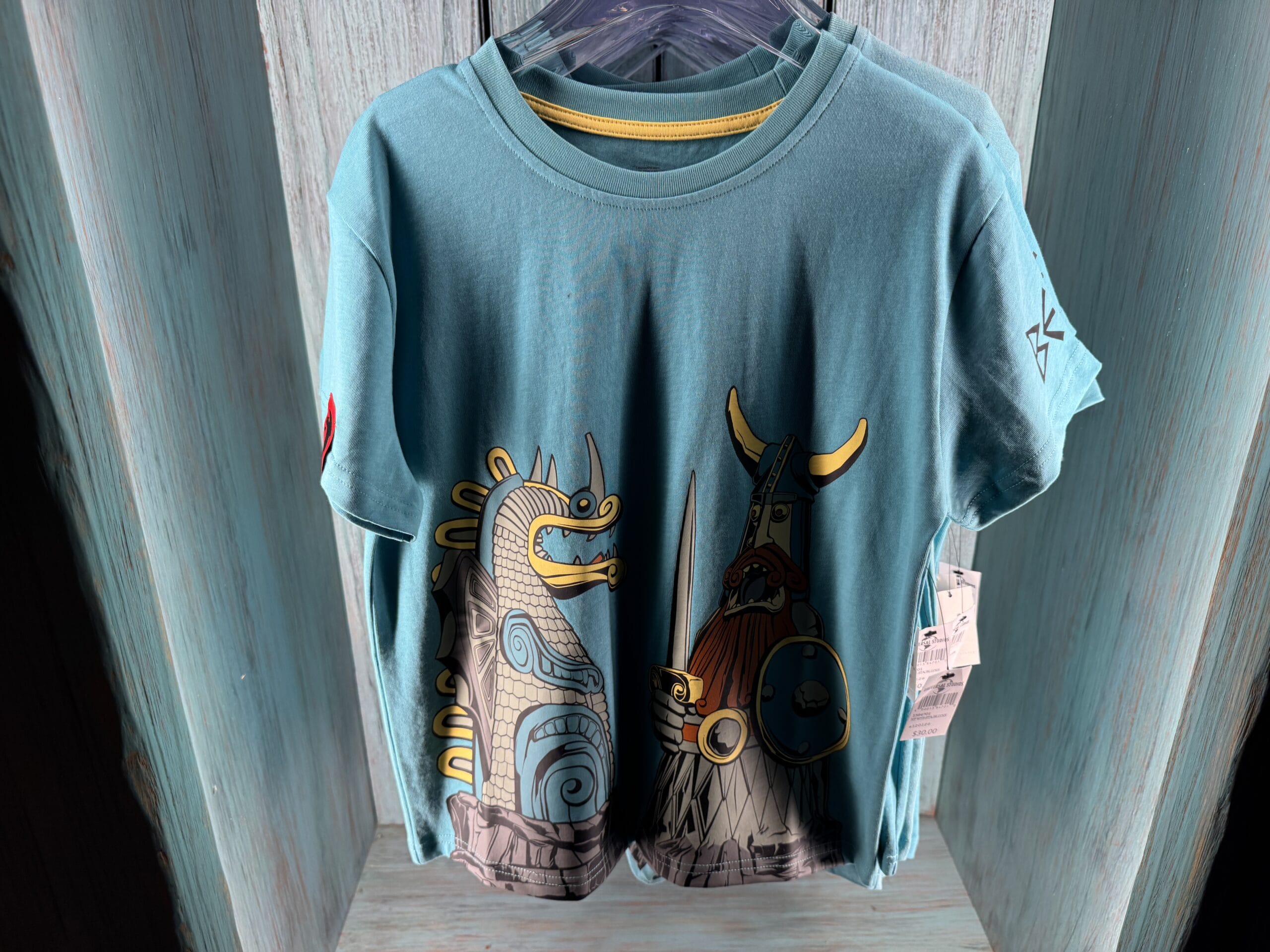 Teal Spit Fyre t-shirt with bright Viking and dragon art on a merch rack, like kiosk stands seen at Disney parks worldwide.