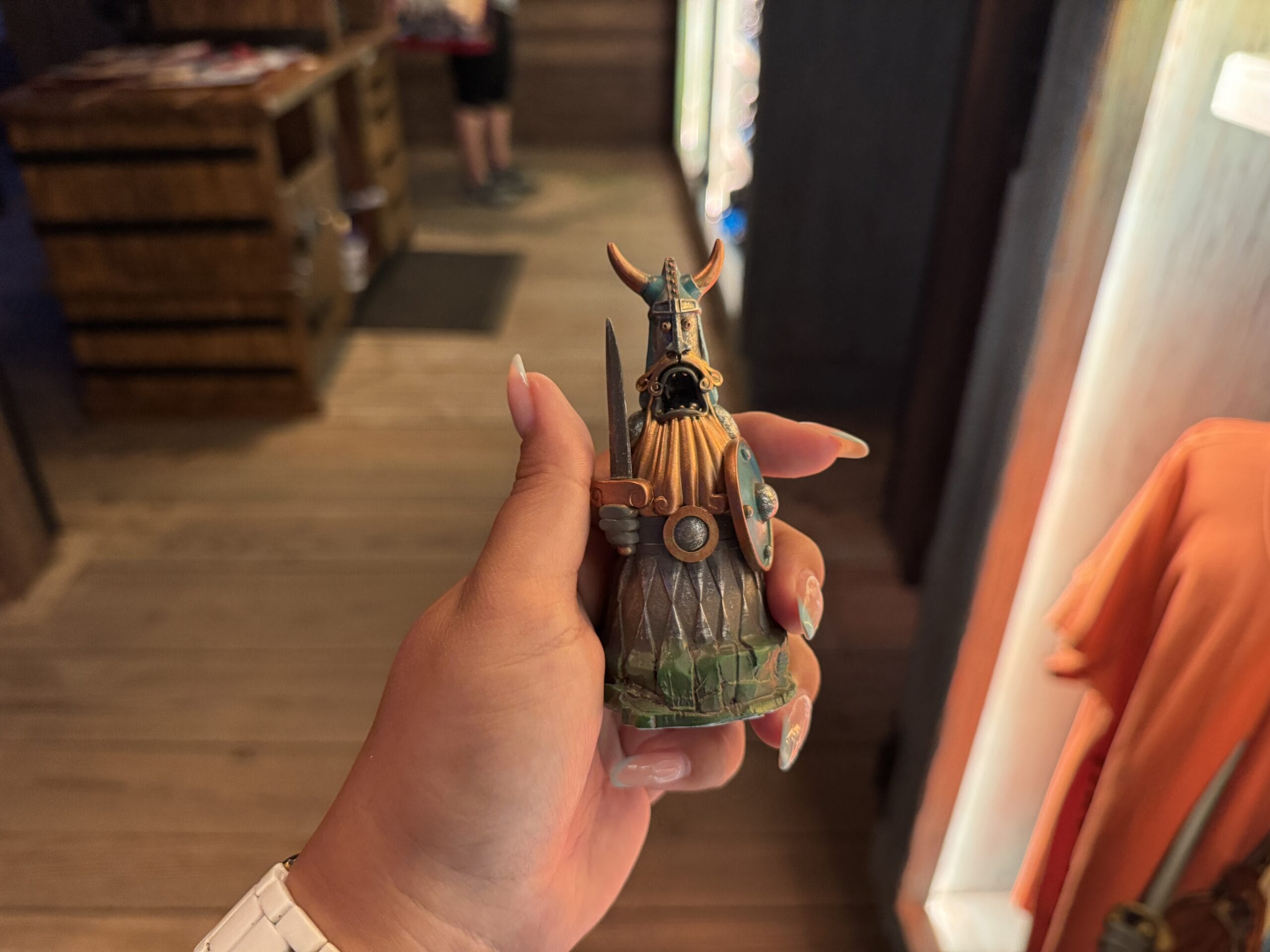 A hand holds a small Viking figurine at the Spit Fyre kiosk, similar in style to Disney park gift shops with themed wooden decor.