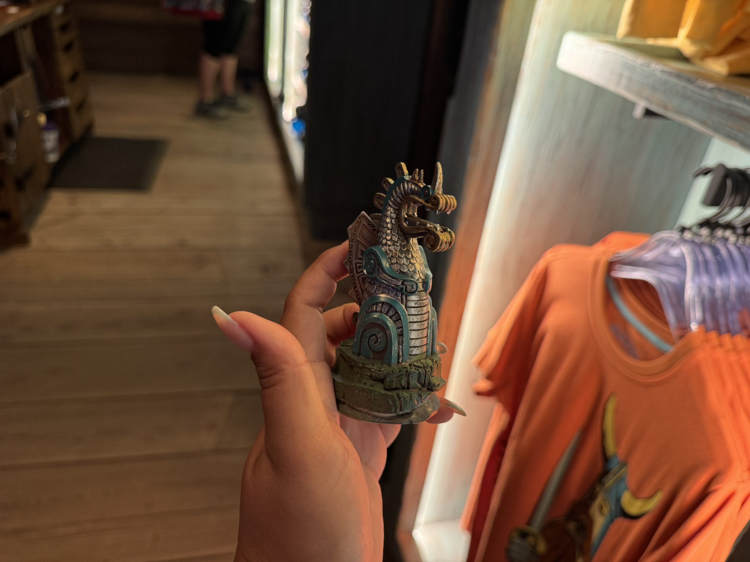 A guest holds a seahorse figurine at the Spit Fyre kiosk, surrounded by vibrant orange shirts, similar to Disney park merchandise.
