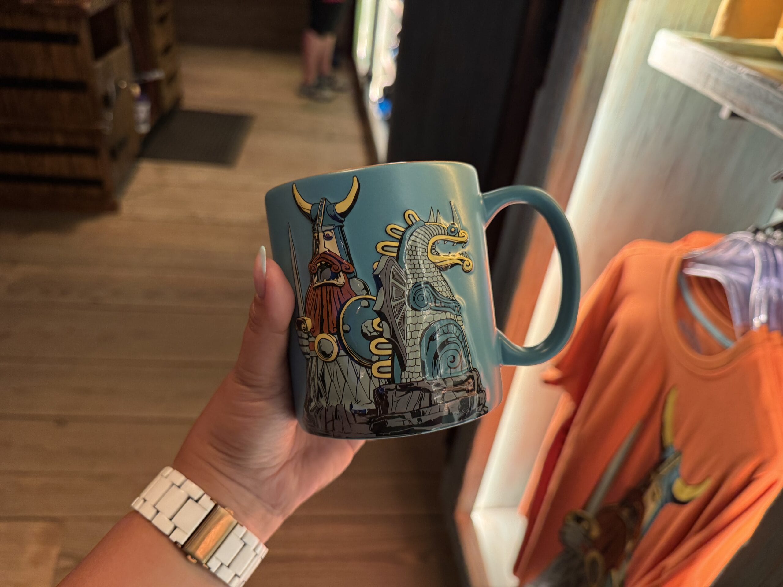A hand holds Spit Fyre merch—a blue mug with Viking ships—orange shirts hang behind, reminiscent of Disney park kiosks.
