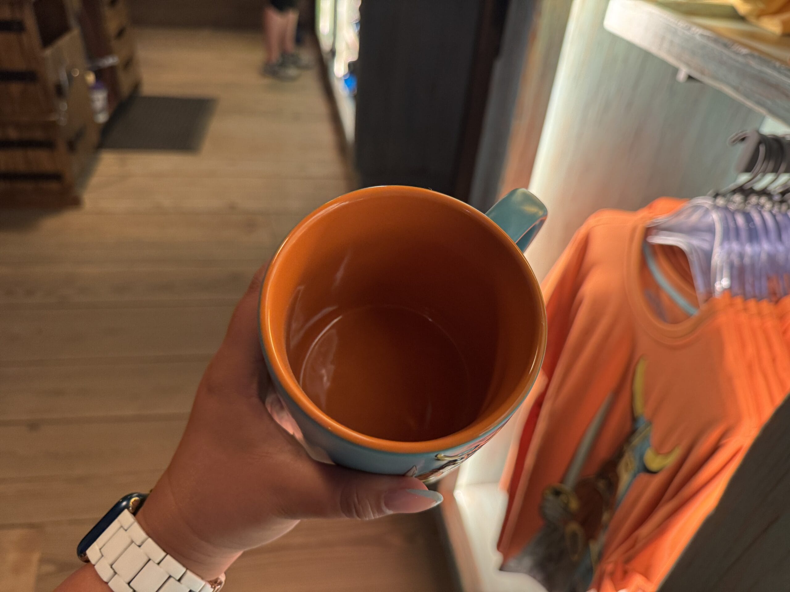 A hand holds an orange Spit Fyre mug inside a Disney park shop, near a kiosk of orange shirts and themed merchandise.