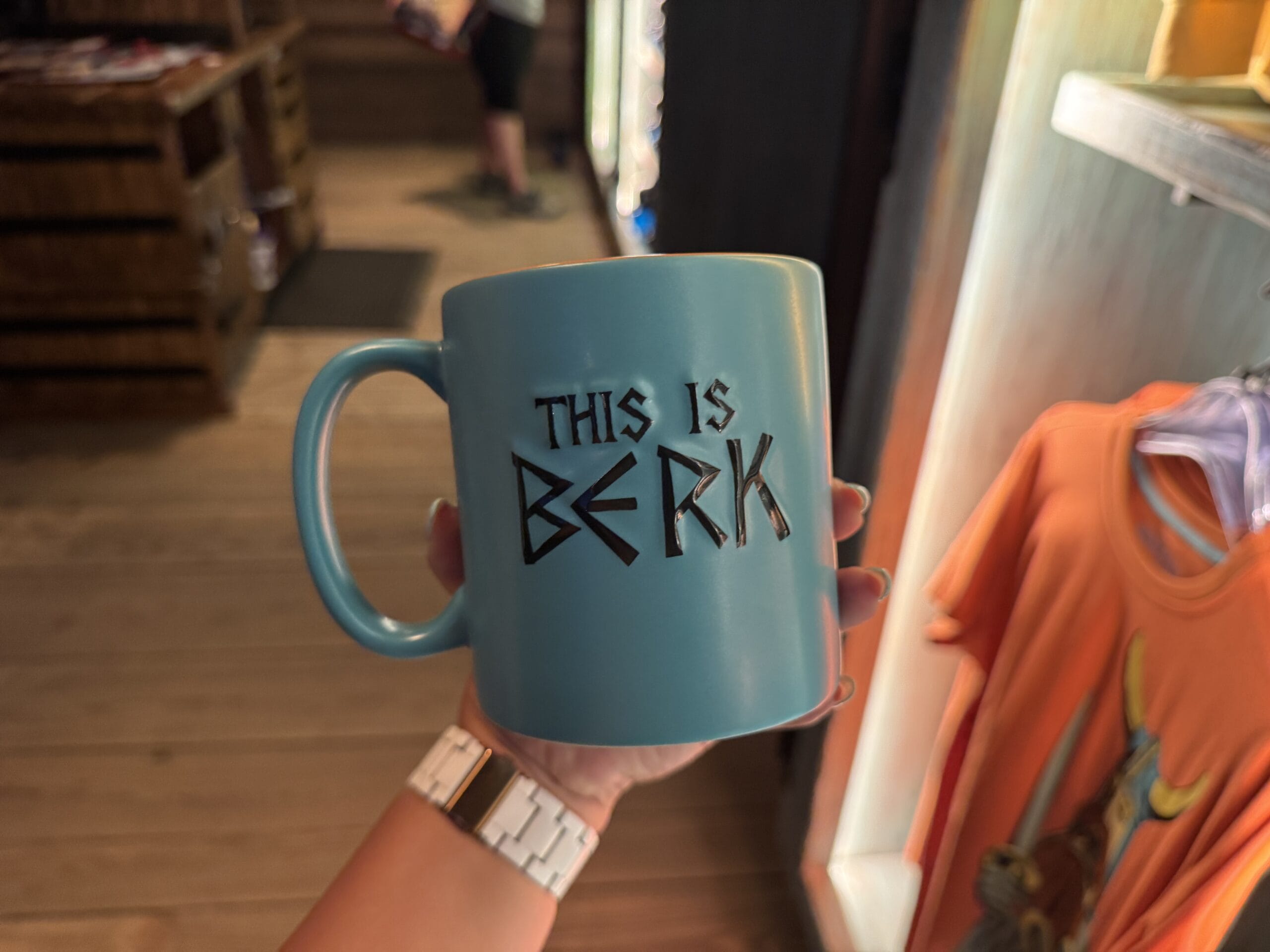 A hand holds Spit Fyre merch: a blue mug reading "THIS IS BERK" in bold, black letters, reminiscent of souvenir mugs at Disney parks.