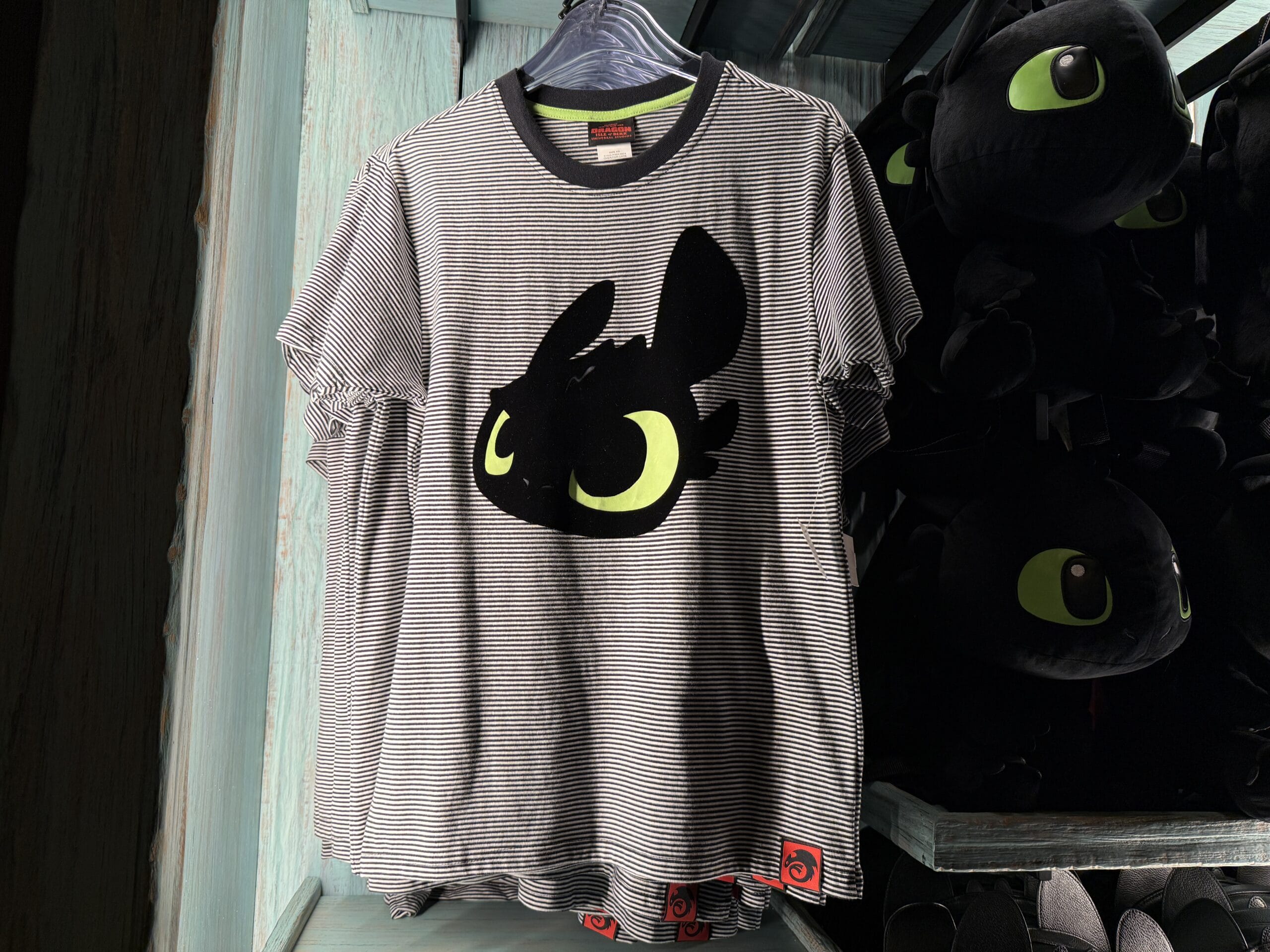 Striped Toothless T-shirt with Spit Fyre graphic on a merch kiosk hanger at a park, plushies in back—castle not pictured.