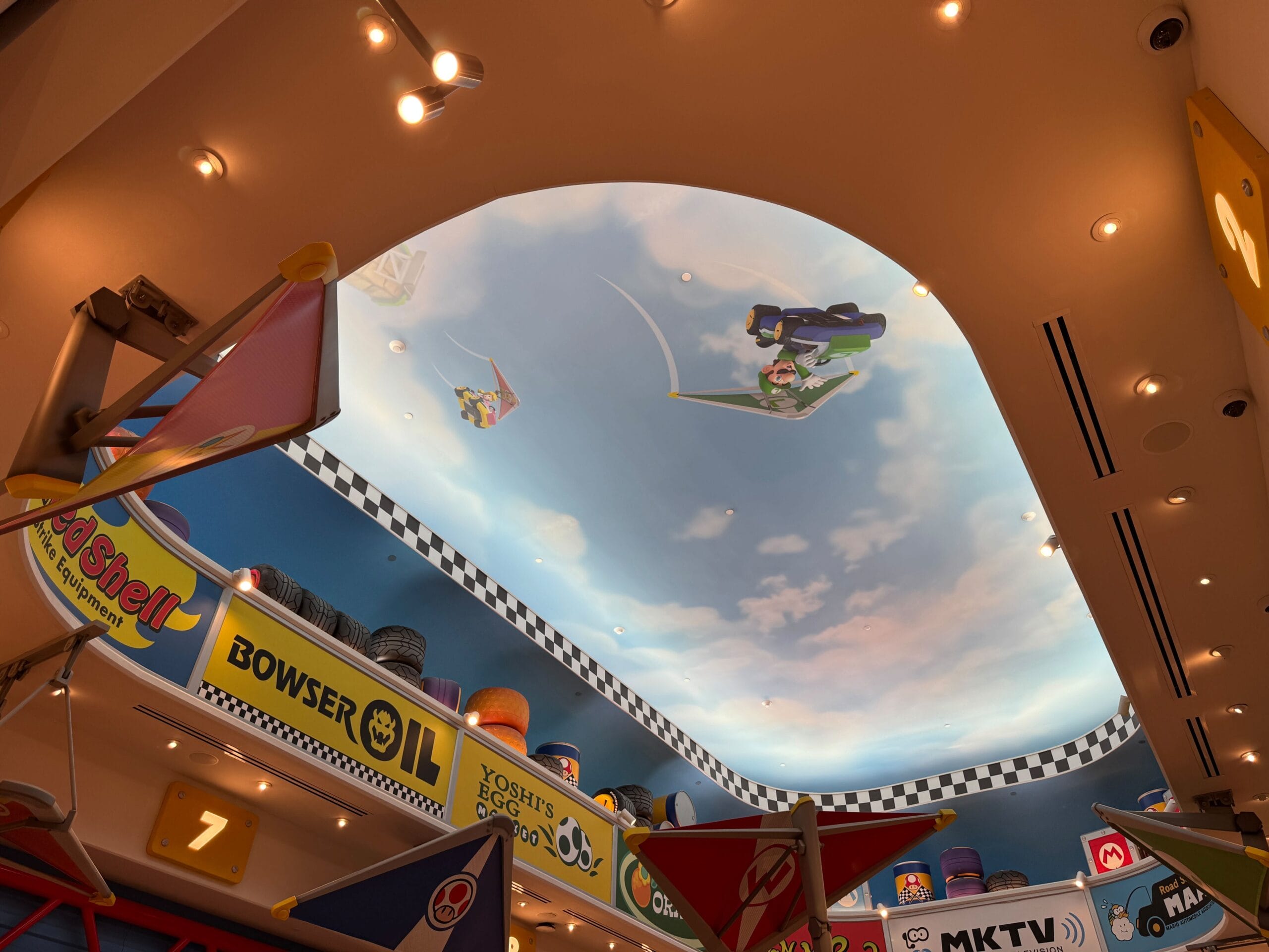 Mario Kart-inspired ceiling mural with airborne racers and race banners, styled like immersive Disney park attractions indoors.