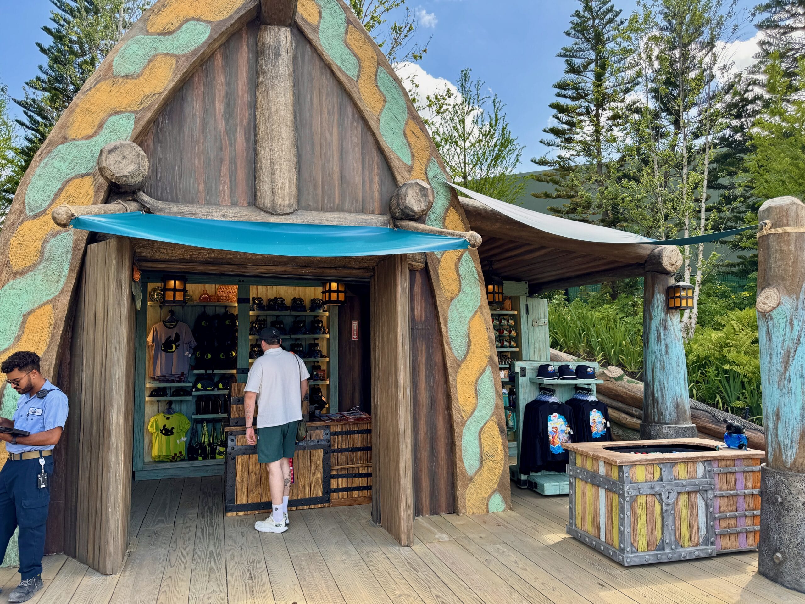 A visitor browses Spit Fyre merchandise in a cozy, wooden kiosk with vibrant trim, reminiscent of Fantasyland shop stalls at Disney Parks.