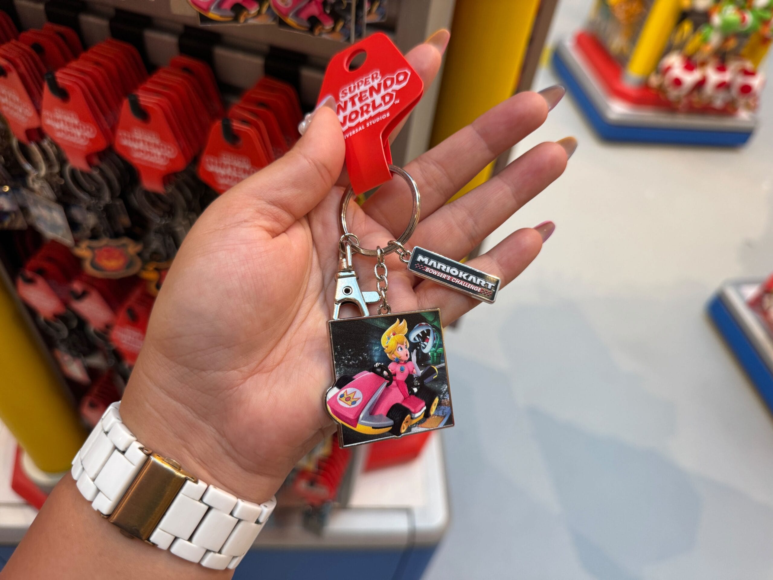 A hand holds Mario Motors merchandise—a Mario Kart keychain with Princess Peach and an Eiffel Tower charm, though not Disney castle related.