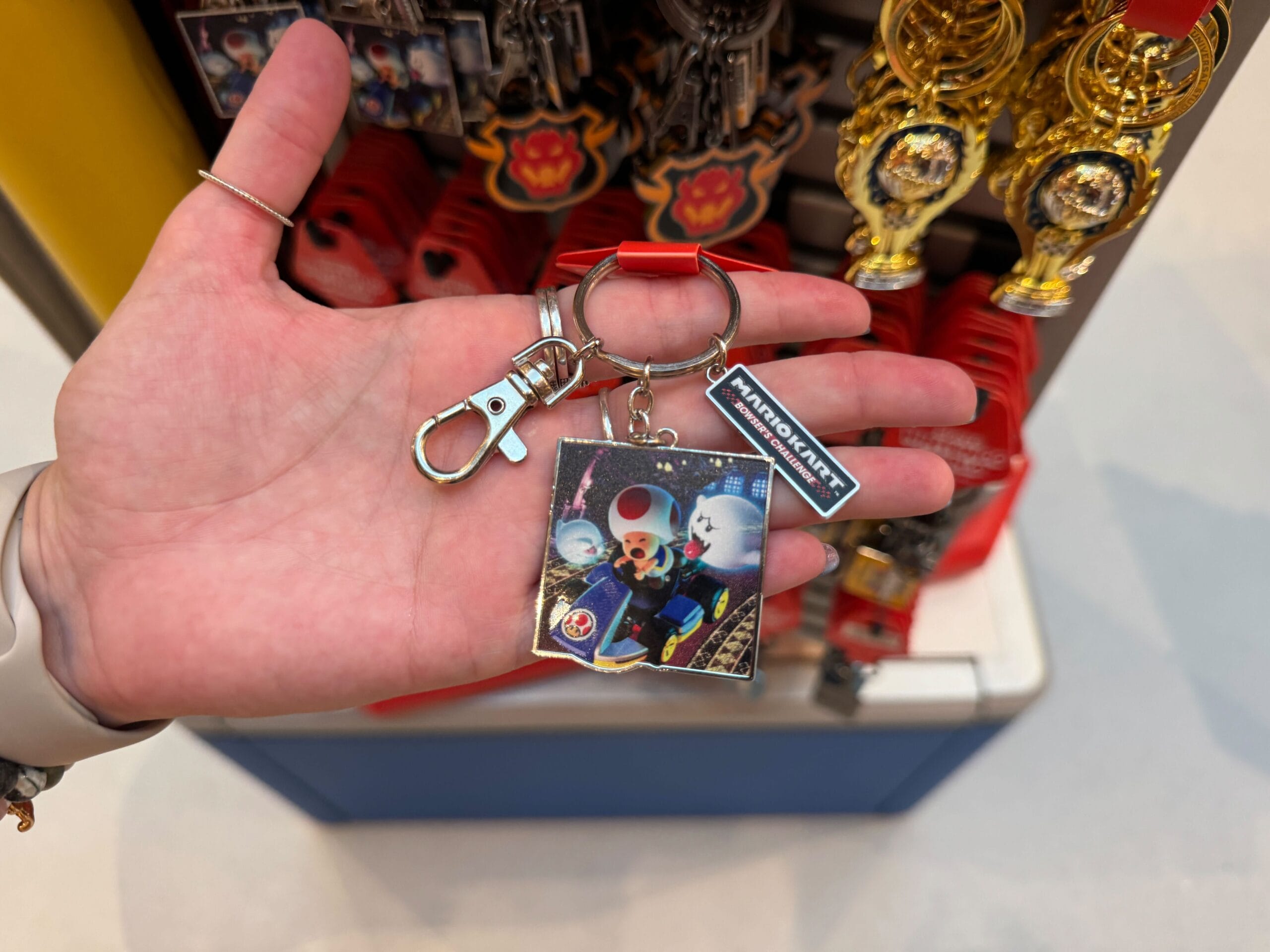 A hand holds a Mario Kart-themed Toad keychain with Mario Motors merchandise behind it, not featuring any Disney park castle.