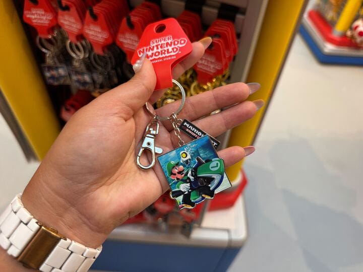 A hand displays a Super Nintendo World Luigi keychain near Mario Motors items; not unlike themed Disney Parks souvenirs, but no castles.