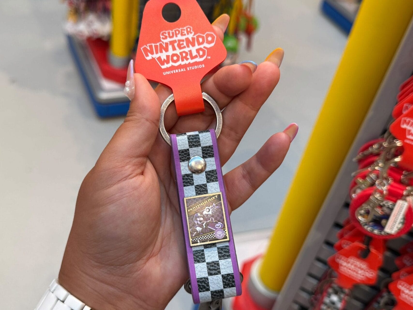 A hand holds a Super Nintendo World keychain with a checkered strap, surrounded by Mario Motors merchandise—nothing like Disney castles.