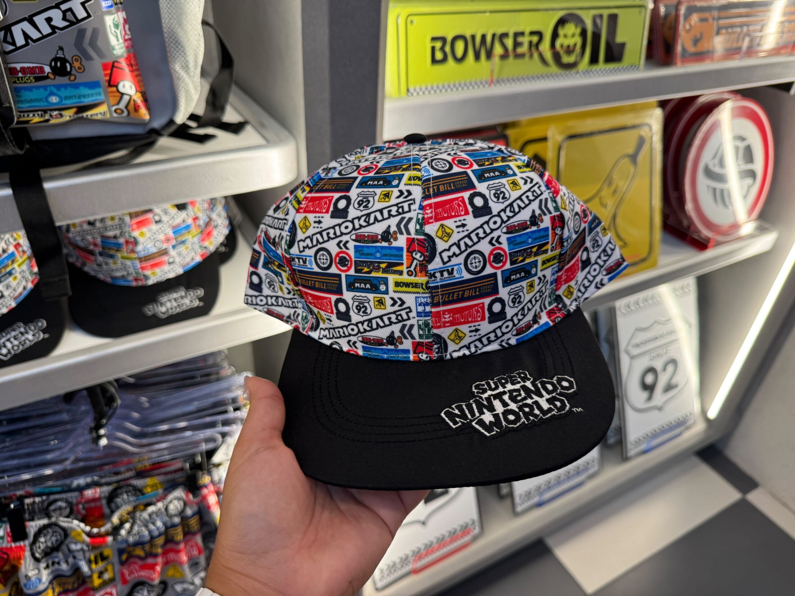 A hand holds a Super Nintendo World Mario Kart hat in a shop with Mario Motors merchandise, reminiscent of immersive theme park stores.