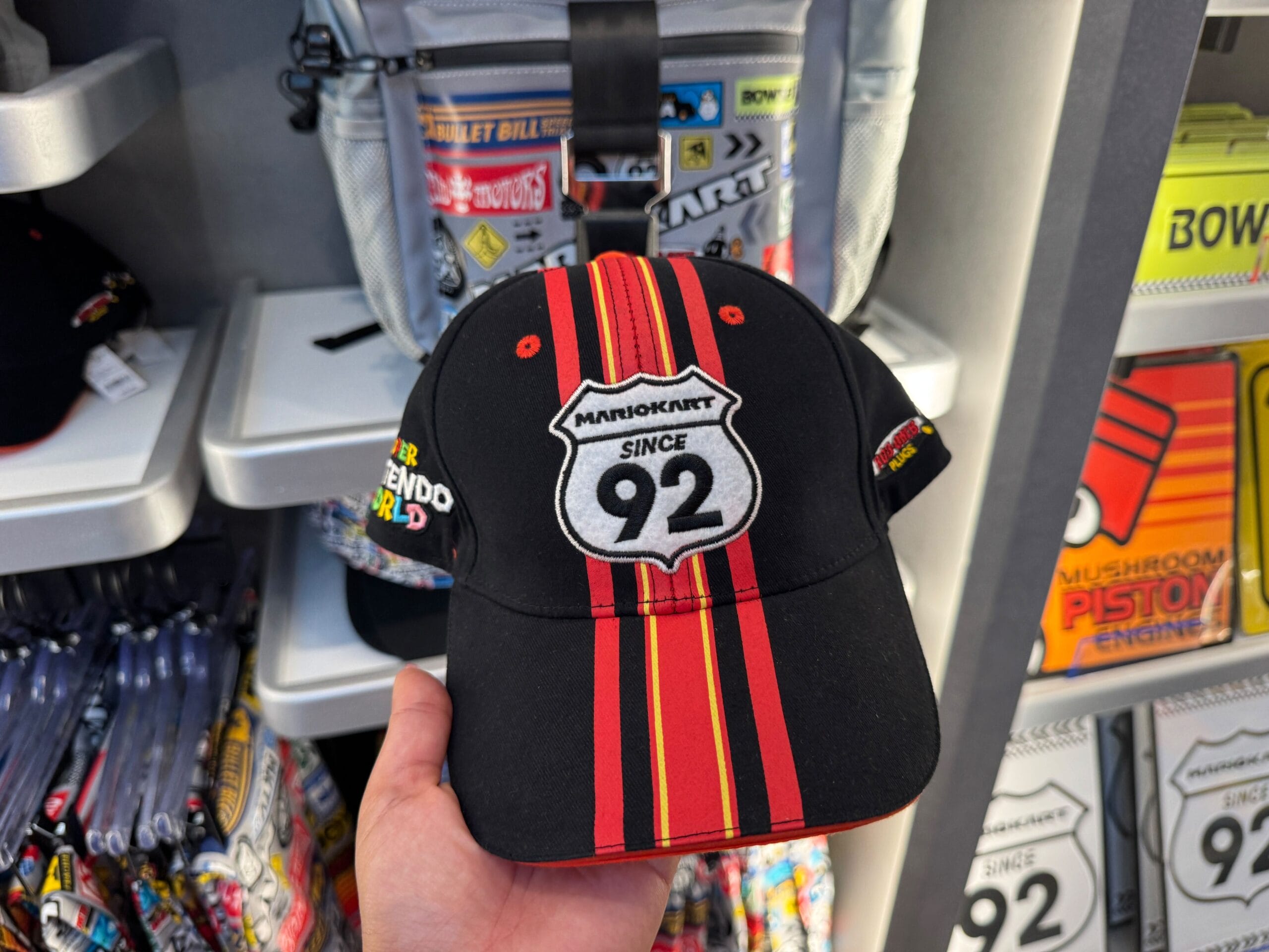 A hand displays Mario Motors gear: a black Mario Kart hat, red and yellow stripes, "Since 92" patch—no Disney castle in view.