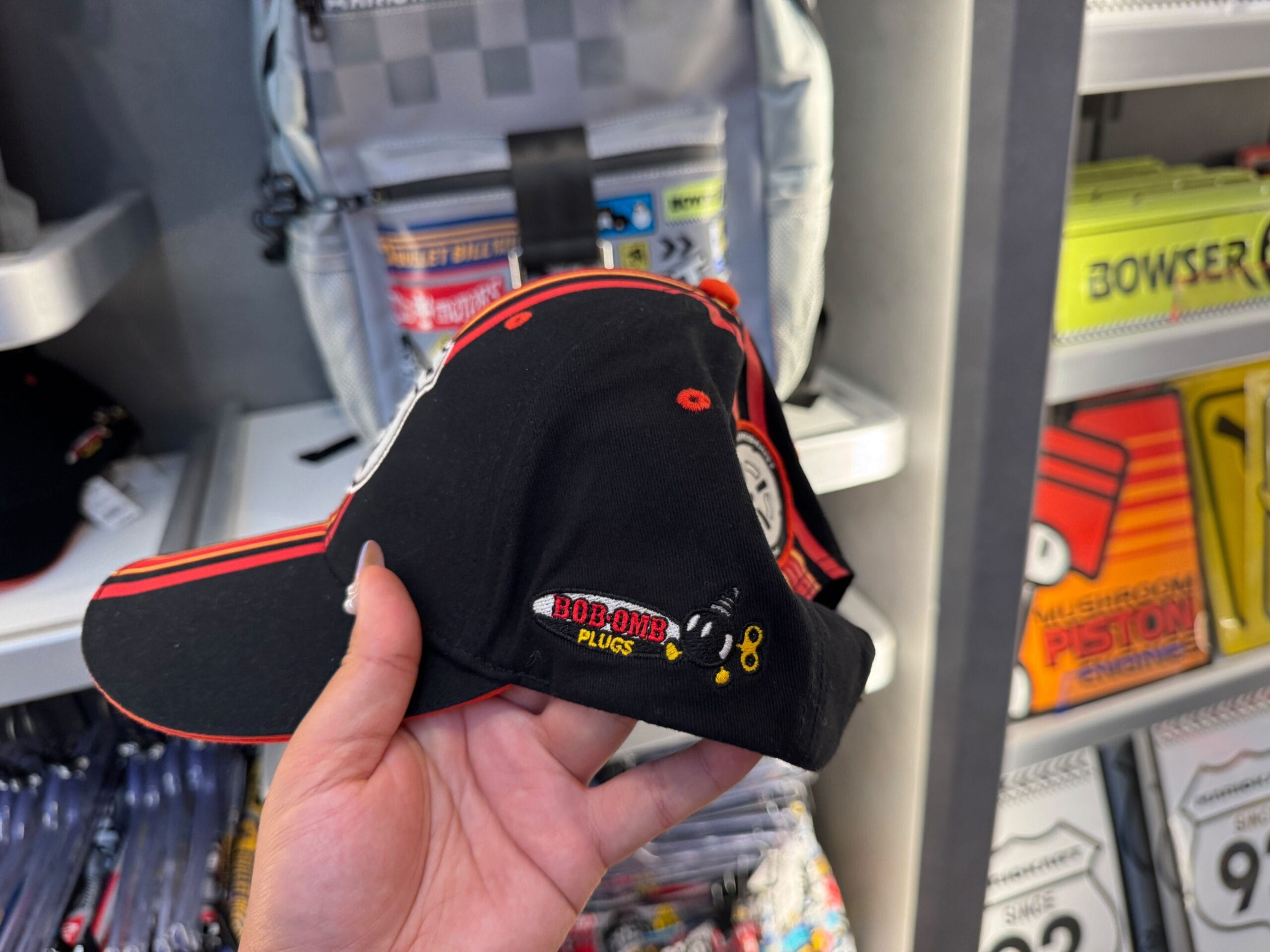 A hand holds a black "Bob-omb Plugs" cap with a cartoon Bob-omb, seen in Super Nintendo World—a playful contrast to Disney castle merch.