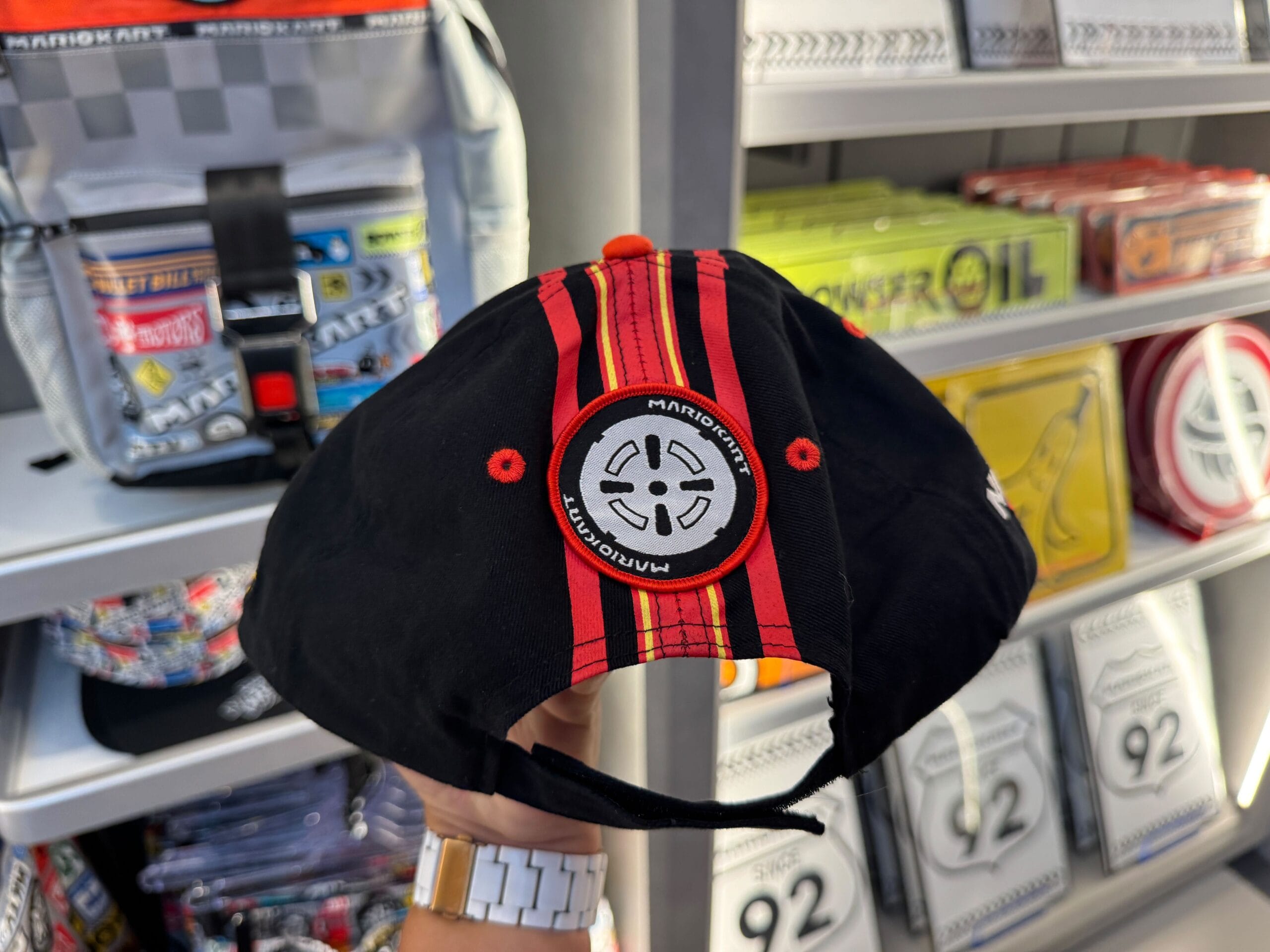 A hand holds a Mario Motors hat with a wheel patch, on display at Super Nintendo World—unlike Disney hat shops near their castles.