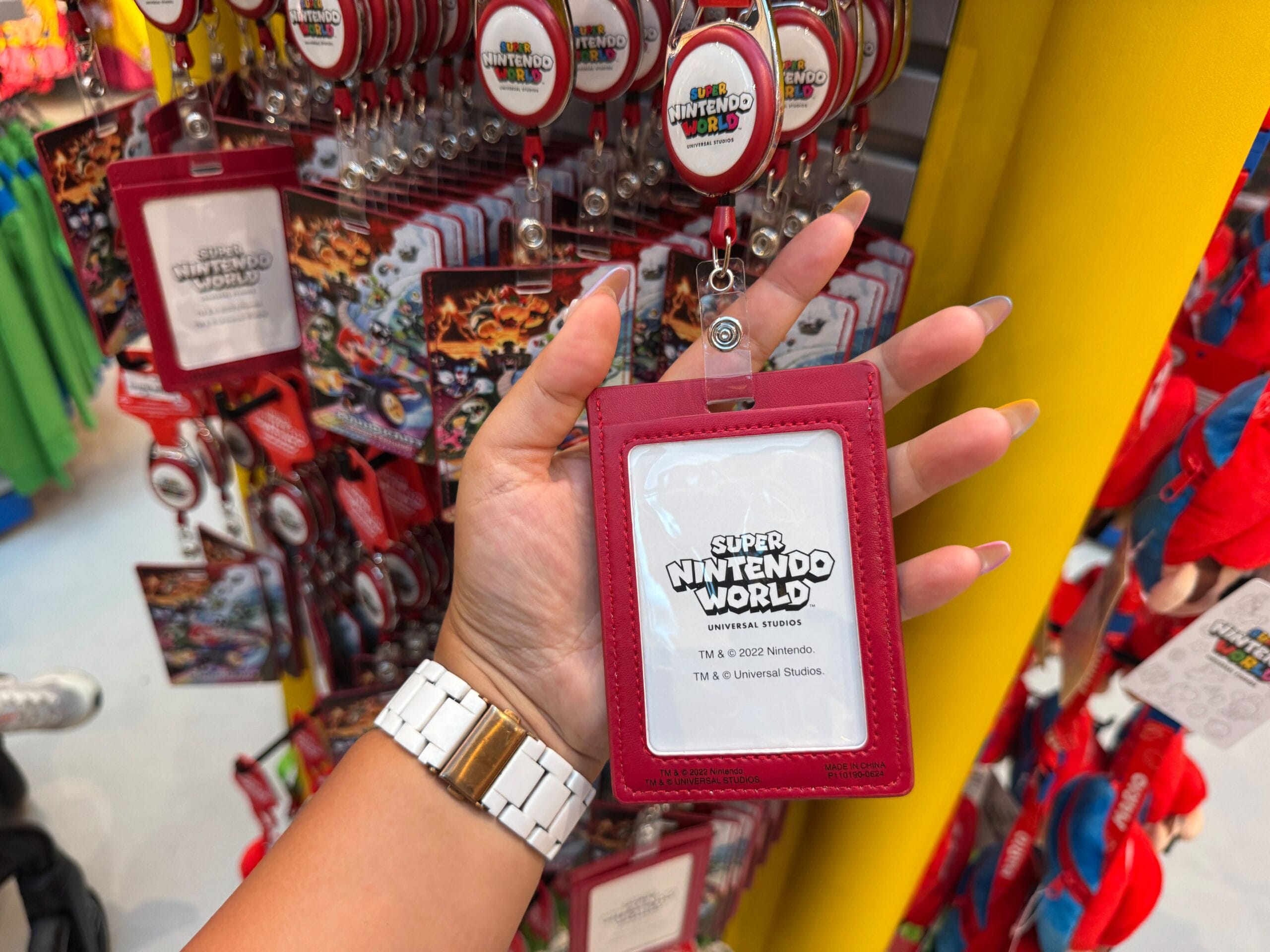 A hand holds a red Super Nintendo World ID holder in front of Mario Motors merchandise, not a Disney castle like at Disney parks.