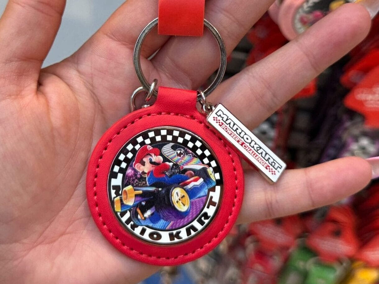 Hand holding a Mario Kart keychain at a themed gift shop, with more keychains behind—unlike Disney parks, no castle in sight.