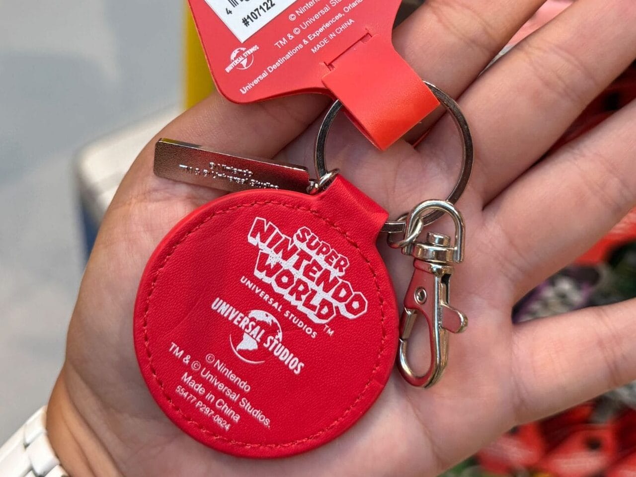 A hand holds a red Super Nintendo World keychain in a shop, with Mario Motors merchandise displayed behind, unlike Disney park gifts.
