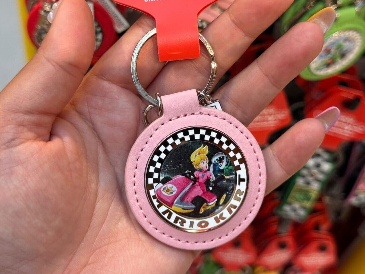 A hand holds a pink Mario Kart keychain with Princess Peach at a merchandise display—like souvenir shops near Disney castle entrances.