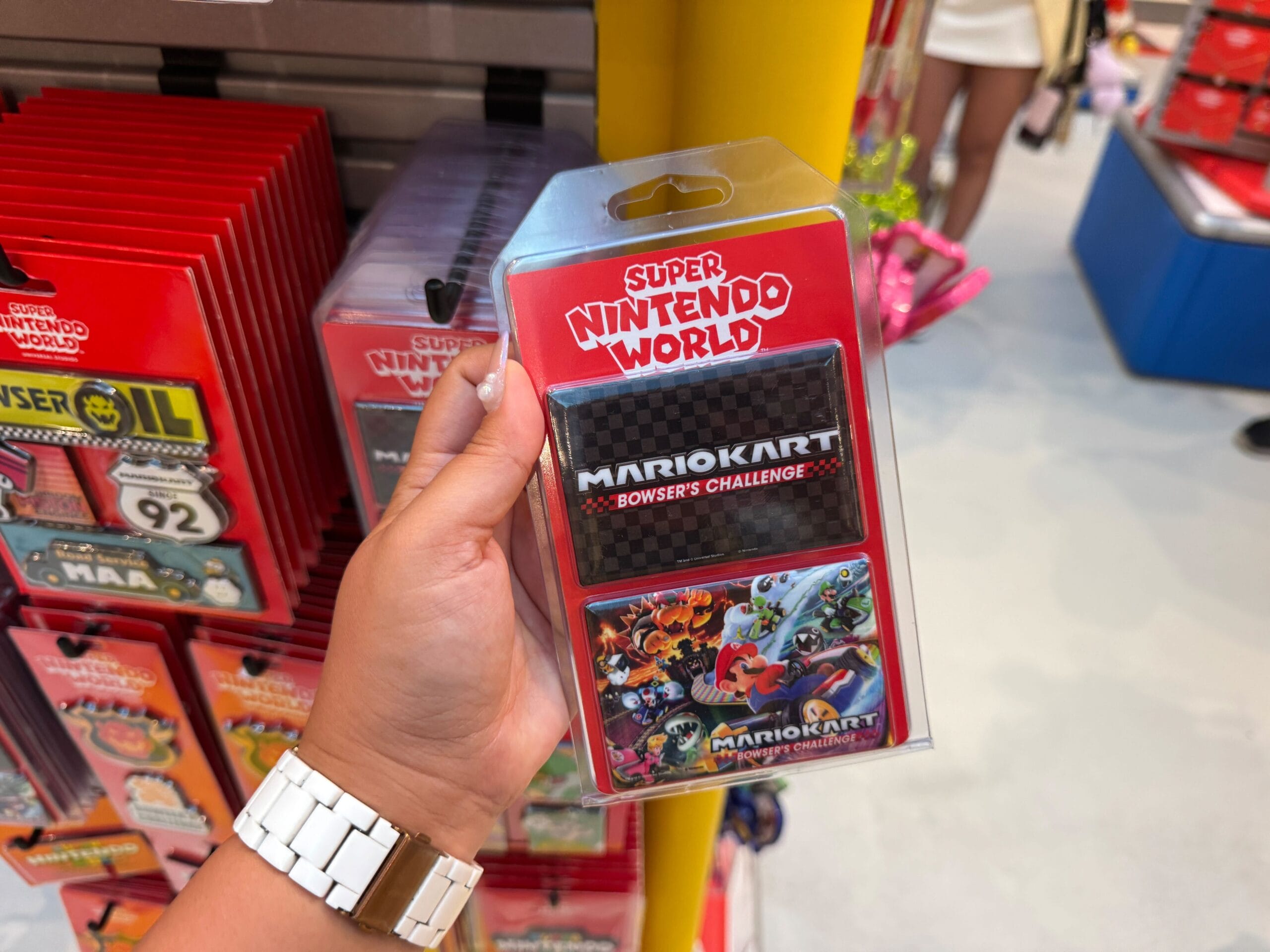 A hand holds a Super Nintendo World Mario Kart: Bowser’s Challenge card pack among themed store merchandise, not Disney castle items.