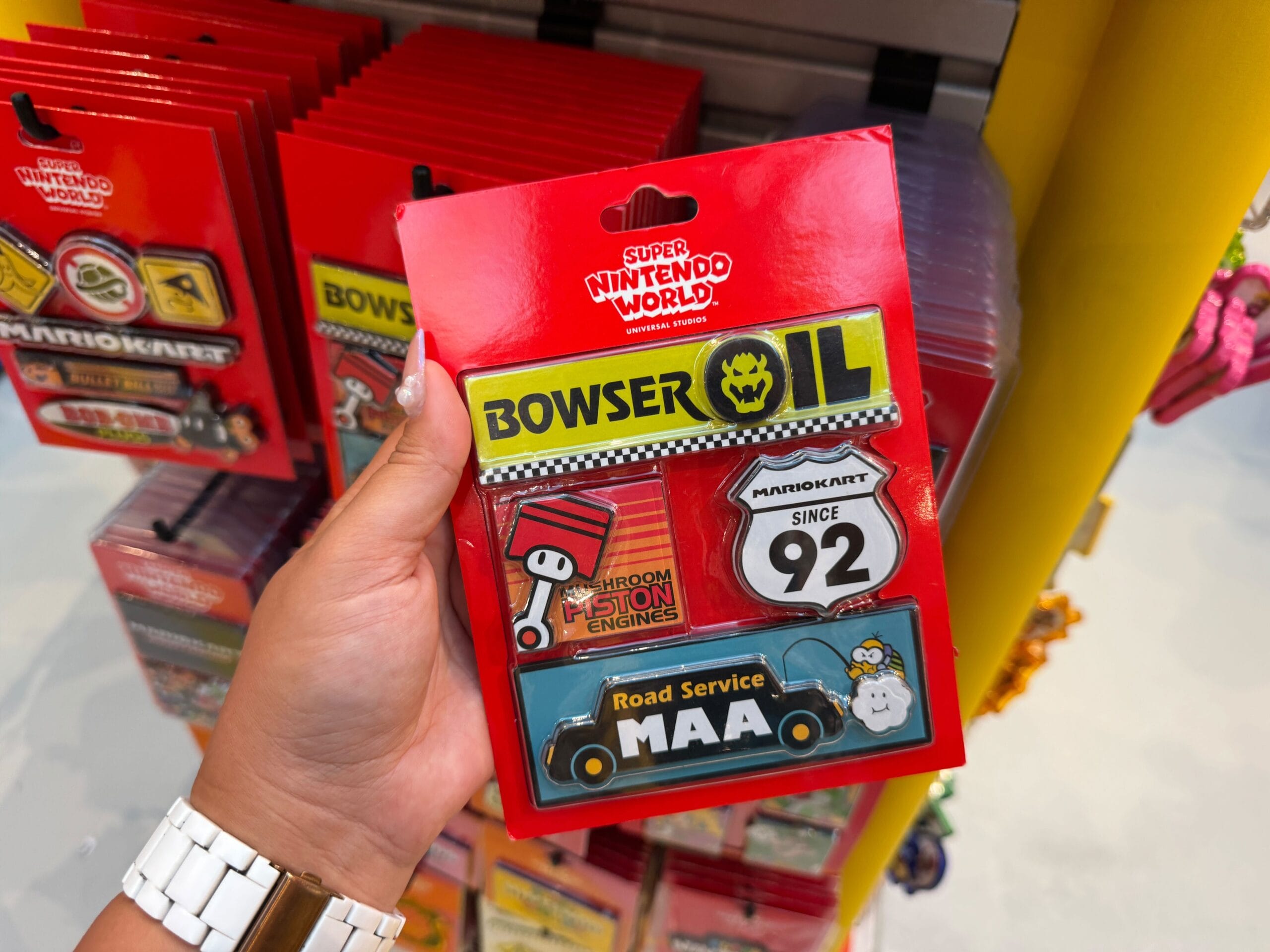 Hand holding Mario Motors pins, like Bowser Oil and Road Service MAA, in Super Nintendo World—no Disney castles like Cinderella’s seen here.