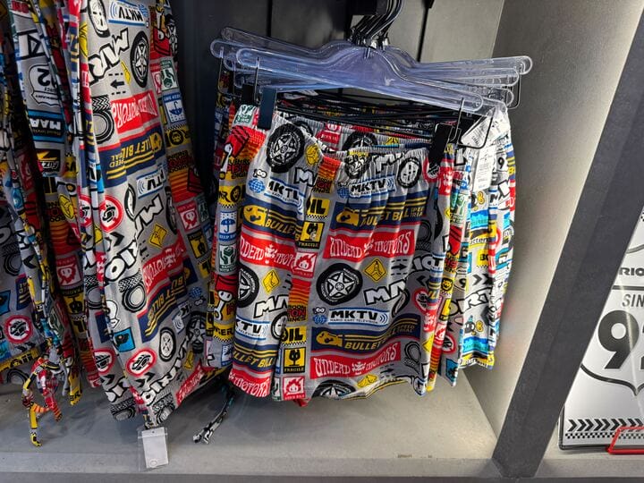 Colorful shorts with bold, graphic Super Nintendo World-style logos hang on a clear hanger in a shop, not Disney Park apparel.