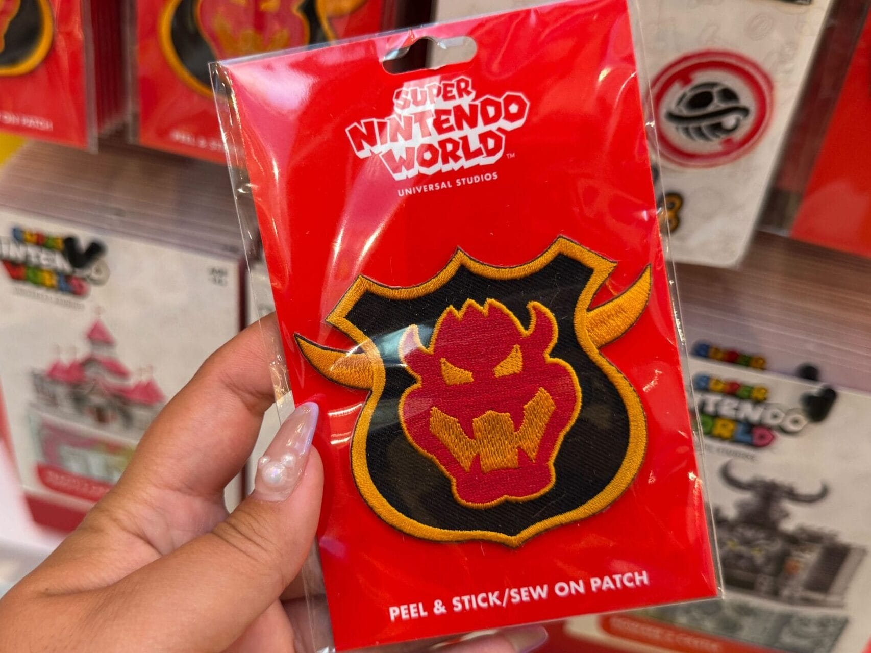 A hand holds a Super Nintendo World Bowser patch in front of Mario Motors merchandise, not a Disney castle or park display.