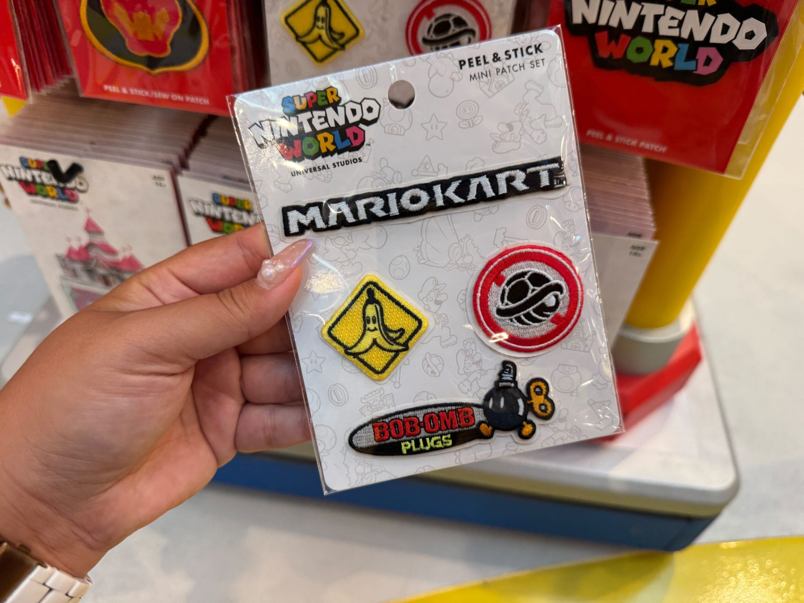 A hand holds a Mario Motors patch set with a banana, shell, and Bob-omb—fun merch like the collectibles seen at Disney Parks.