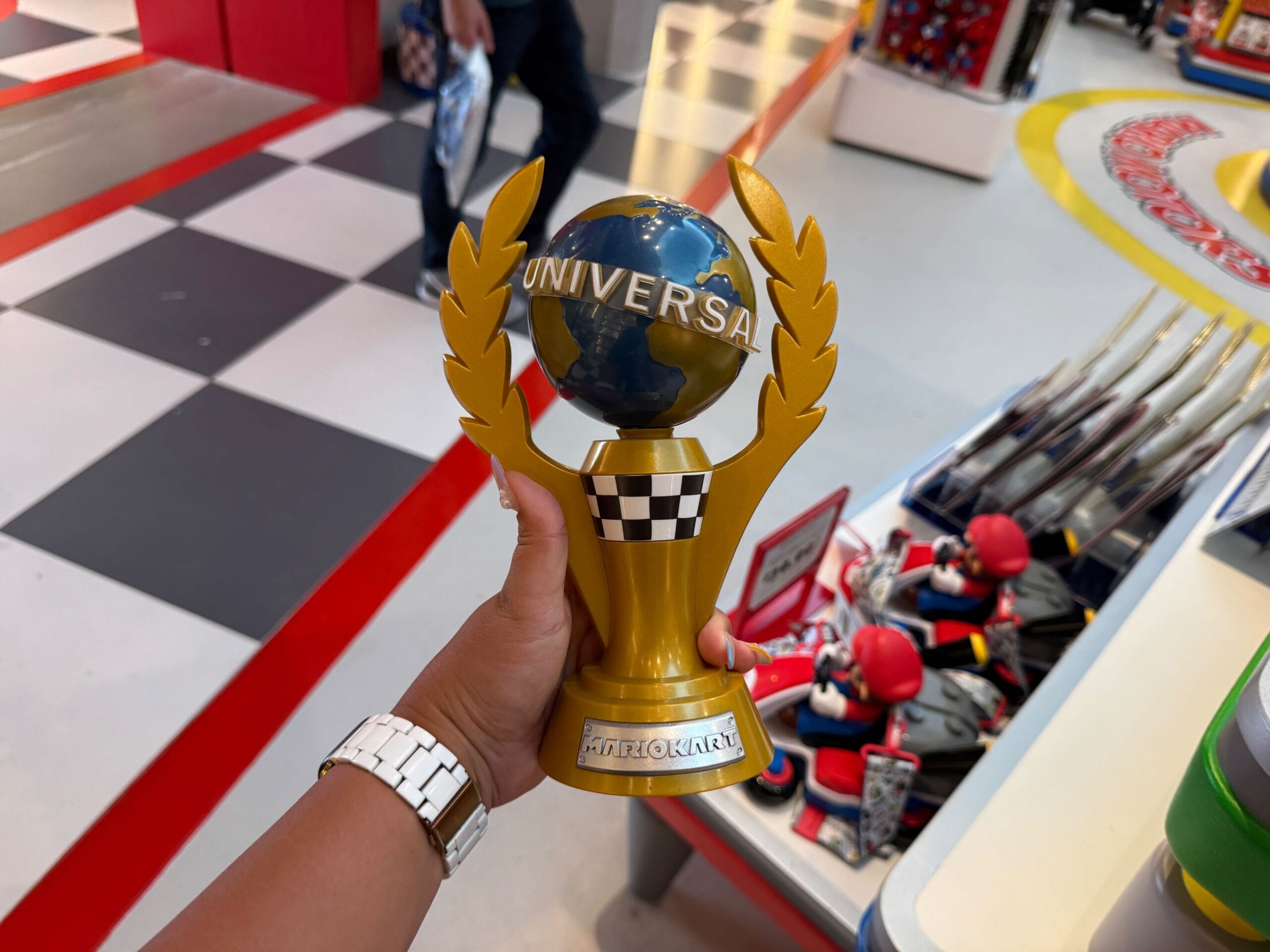 Hand holding a gold Mario Kart trophy in a vibrant Super Nintendo World shop, shelves lined with Mario merchandise—no Disney castle.