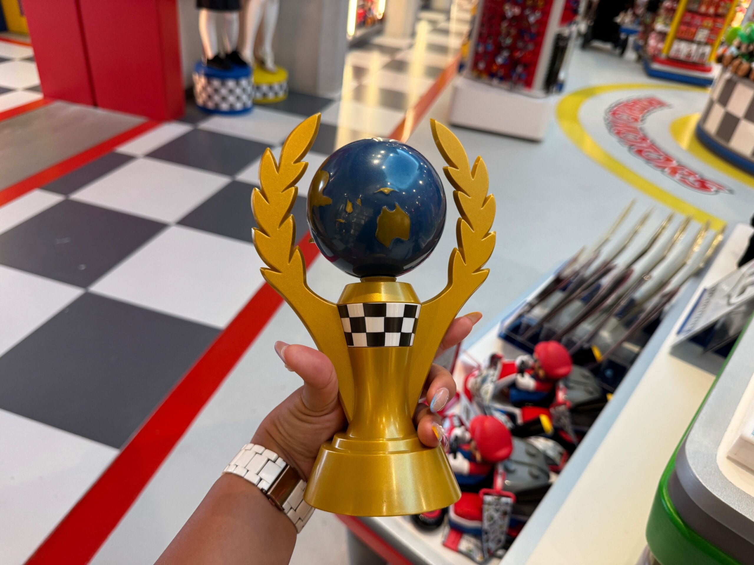 A hand holds a gold trophy with a globe and laurel in a vibrant store with checkered floors, reminiscent of Disney park shops.