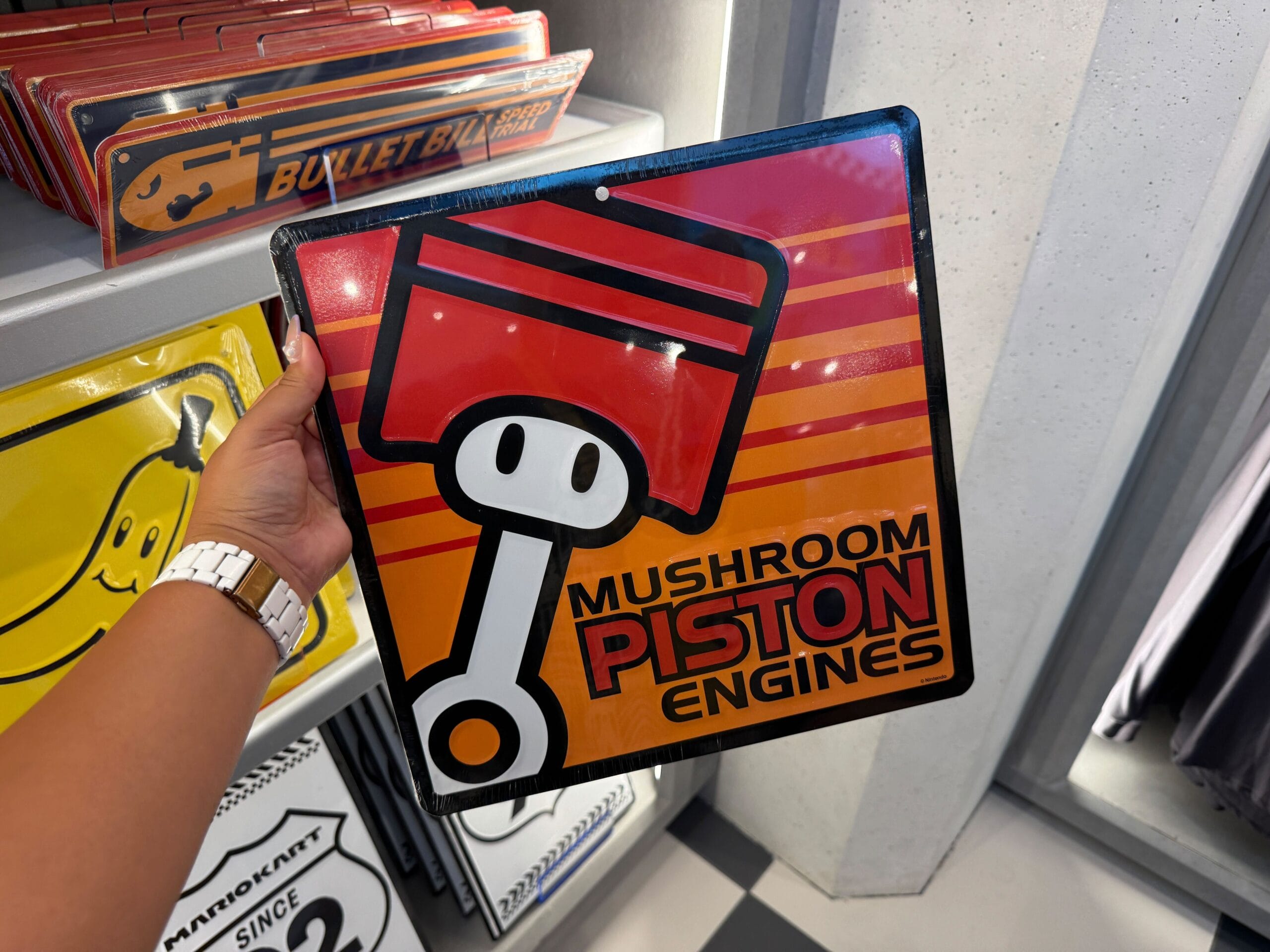 A hand holds a "Mushroom Piston Engines" sign, reminiscent of themed signs at Disney parks but inspired by Super Nintendo World.