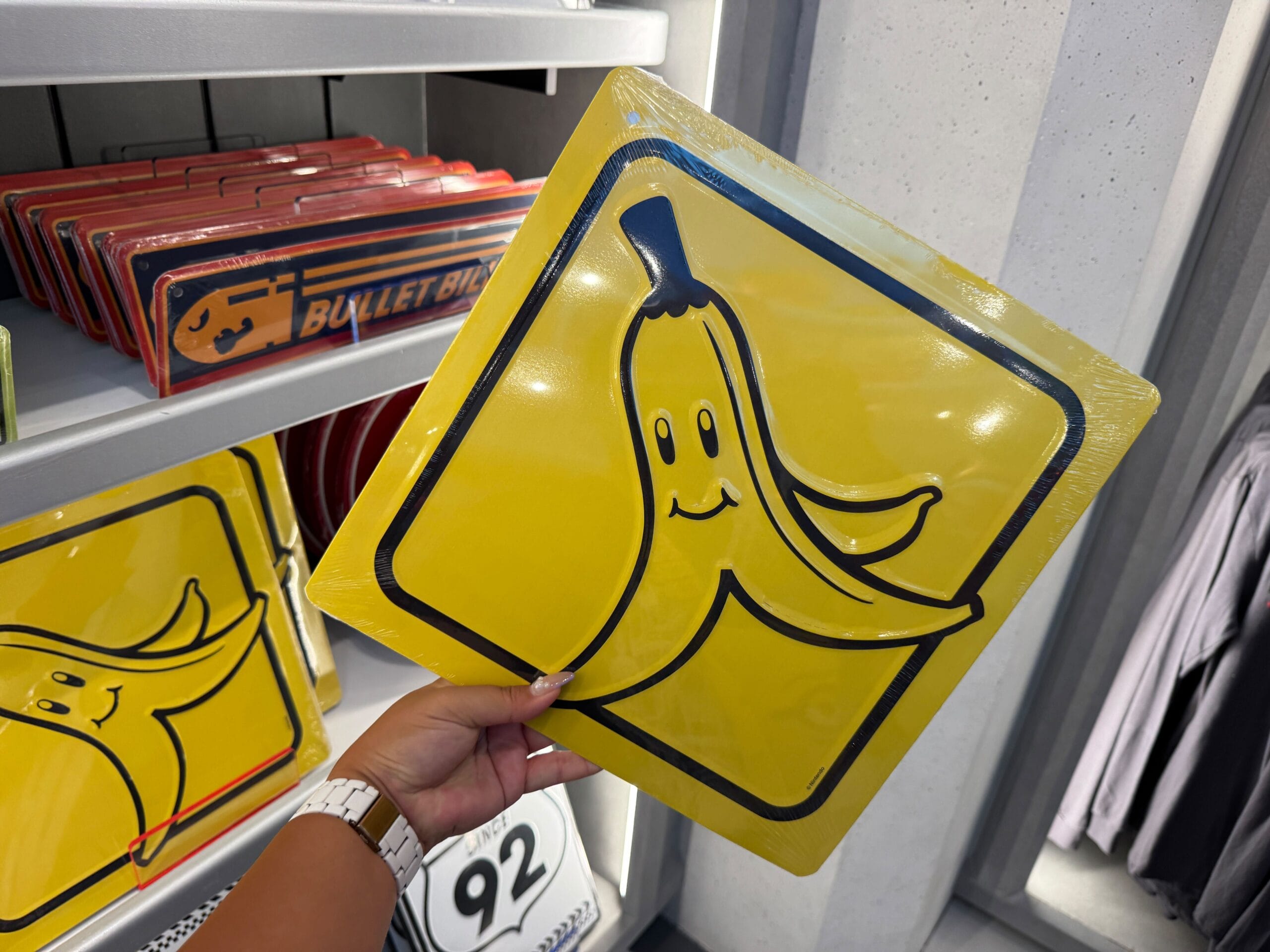 A hand holds a yellow sign with a smiling banana peel inside a themed store, reminiscent of playful Disney park merchandise displays.