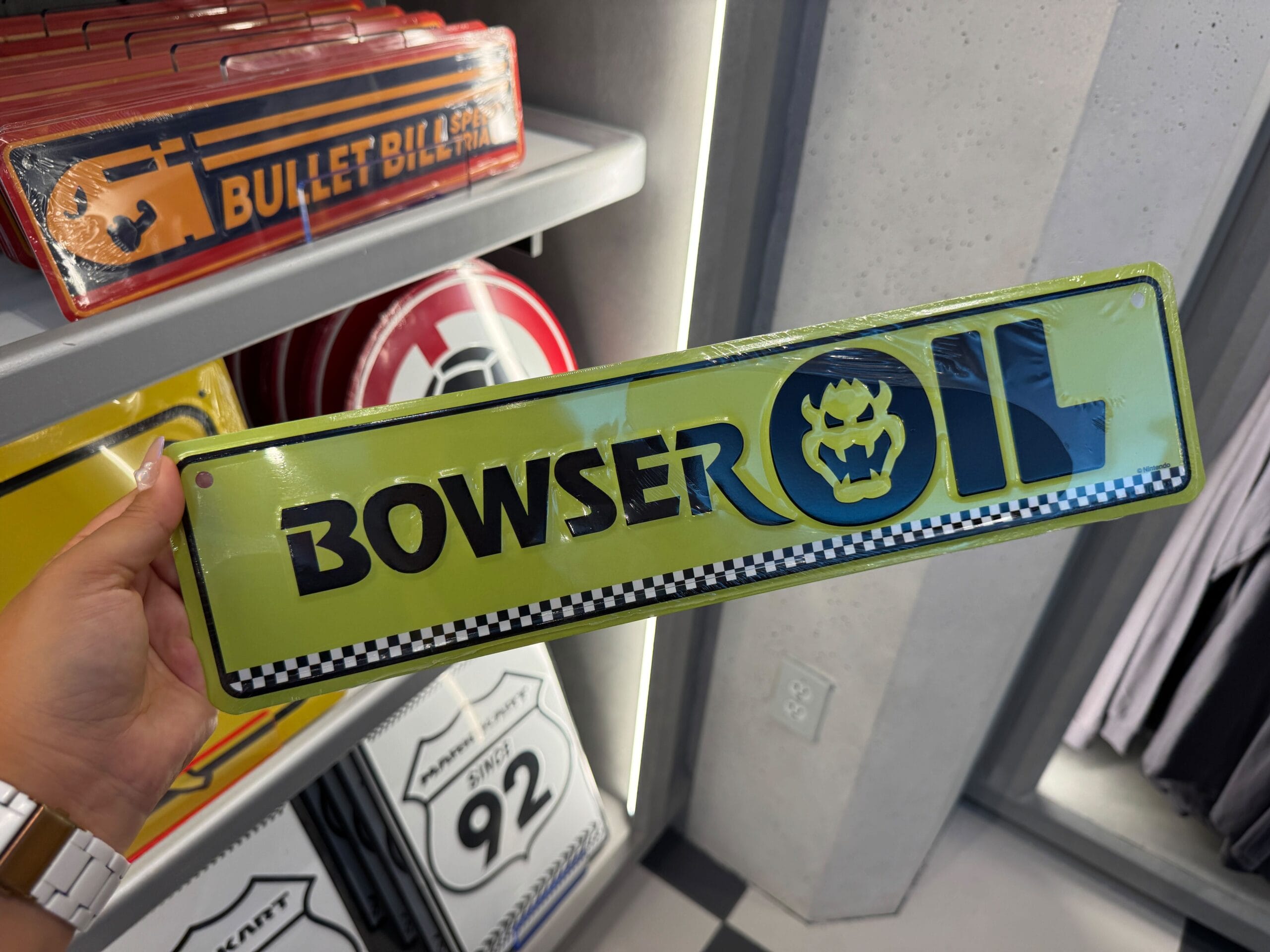 A hand holds a green "Bowser Oil" metal sign—Mario Motors merchandise, reminiscent of playful souvenirs found in Disney Parks.